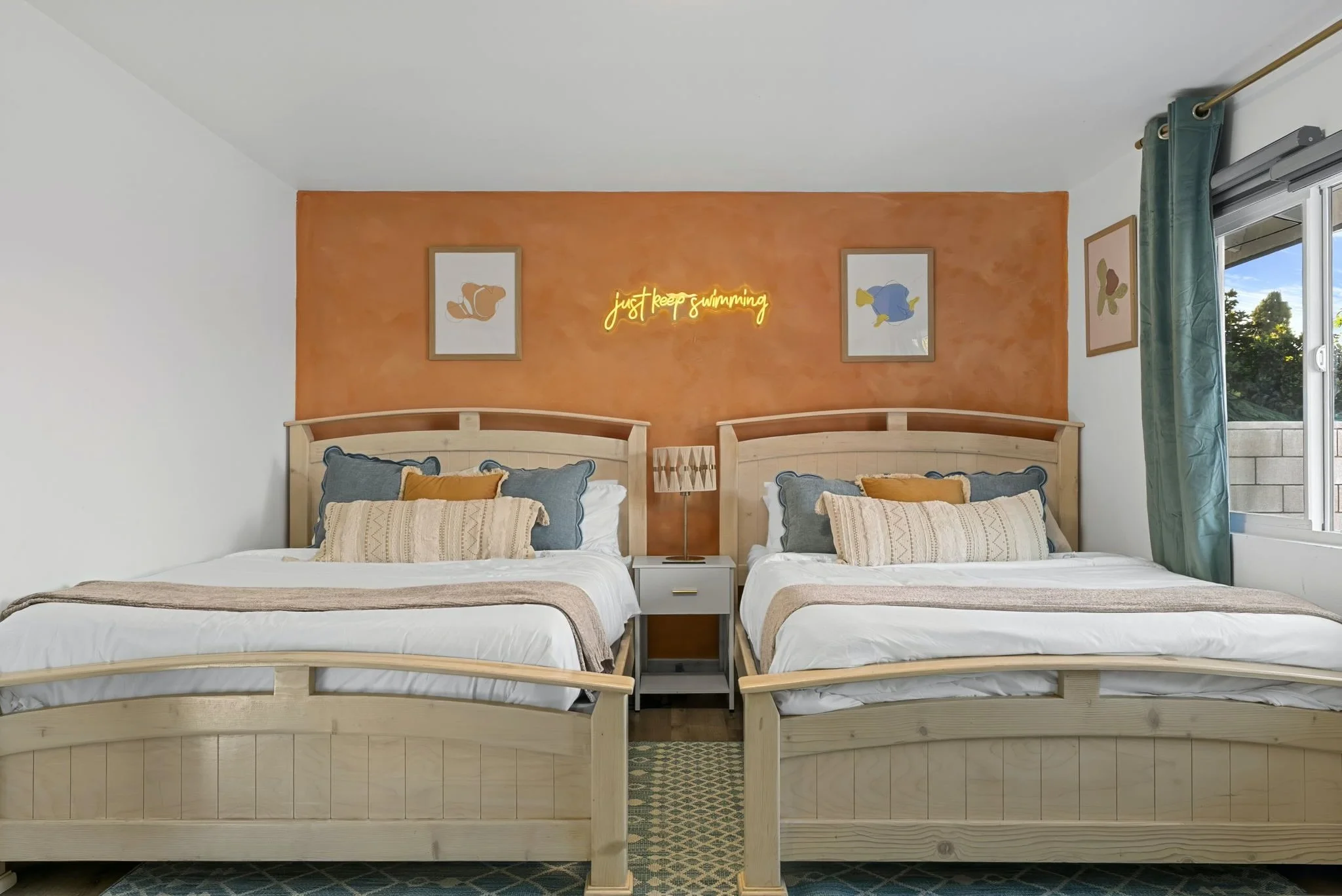 Bright coastal guest bedroom with two twin beds, blue and yellow pillows, orange accent wall, ocean-themed artwork, and a glowing “Just Keep Swimming” sign