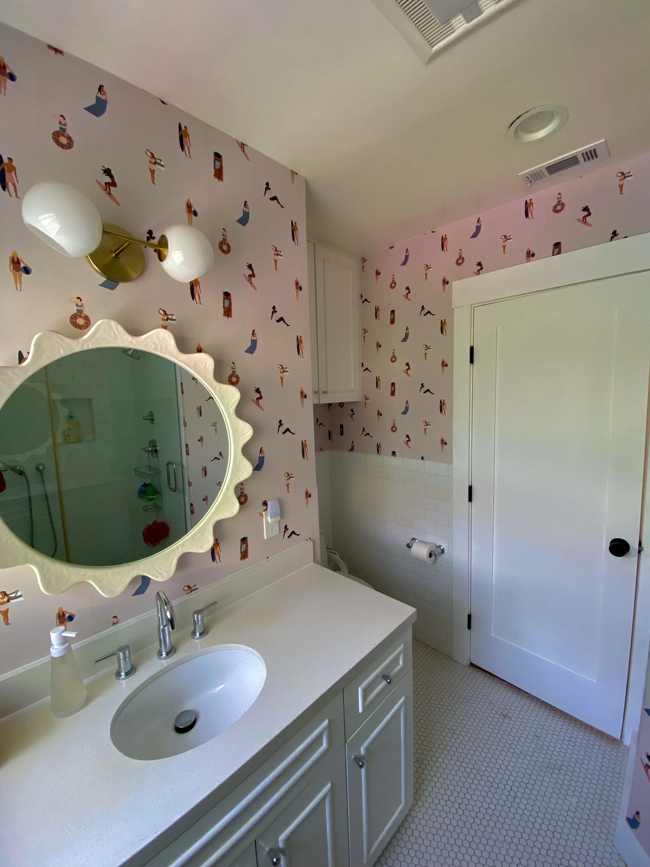 Bright bathroom with pink vintage swimmer-pattern wallpaper, white vanity and sink, scalloped mirror, globe wall light, glass shower, and hexagon tile floor.