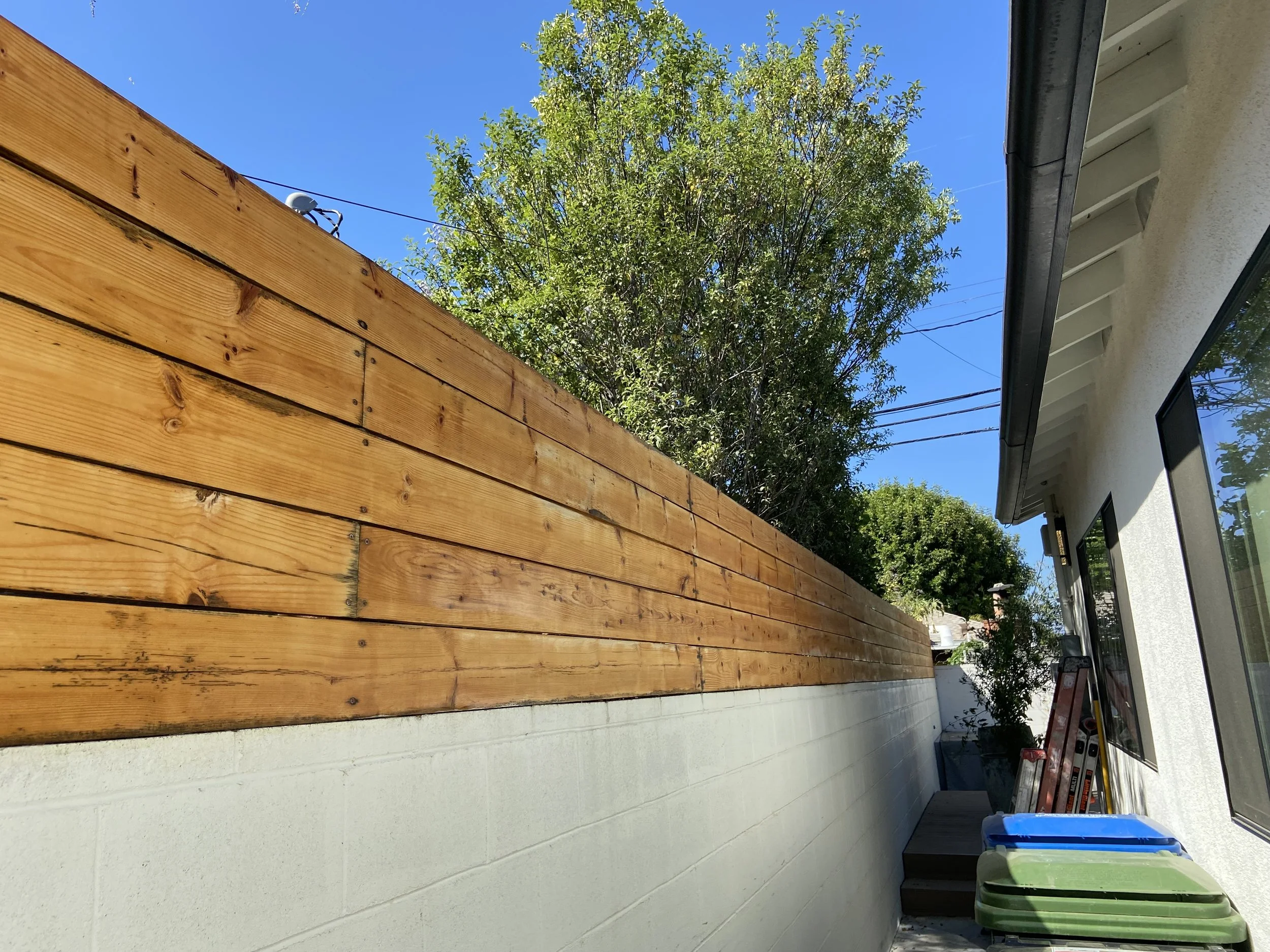 Sunny residential side yard with a horizontal wooden fence, leafy trees, white stucco house, utility wires, ladder, and recycling bins.