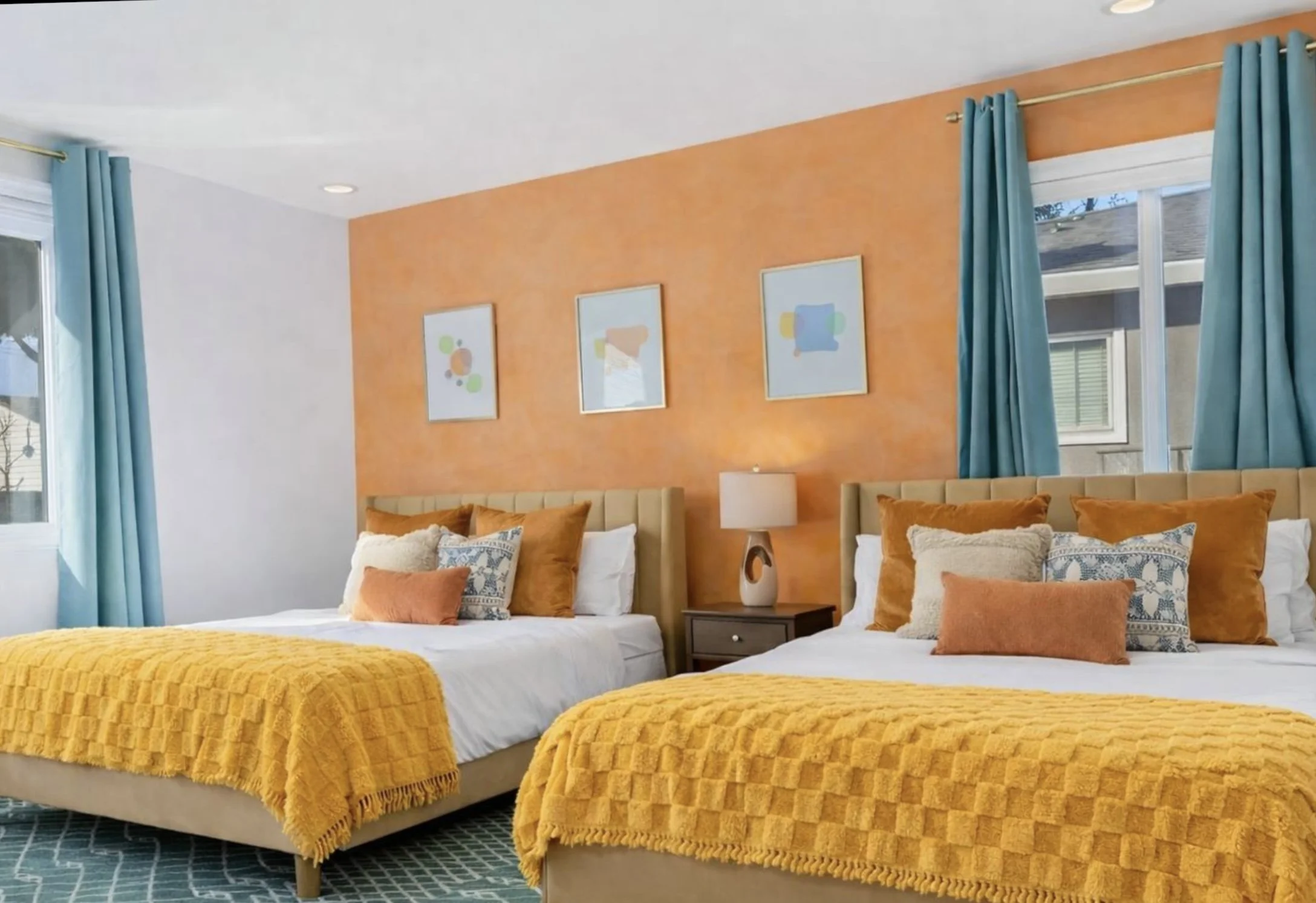 Colorful bedroom with two beds, mustard yellow throws, orange and blue pillows, turquoise curtains, and abstract wall art