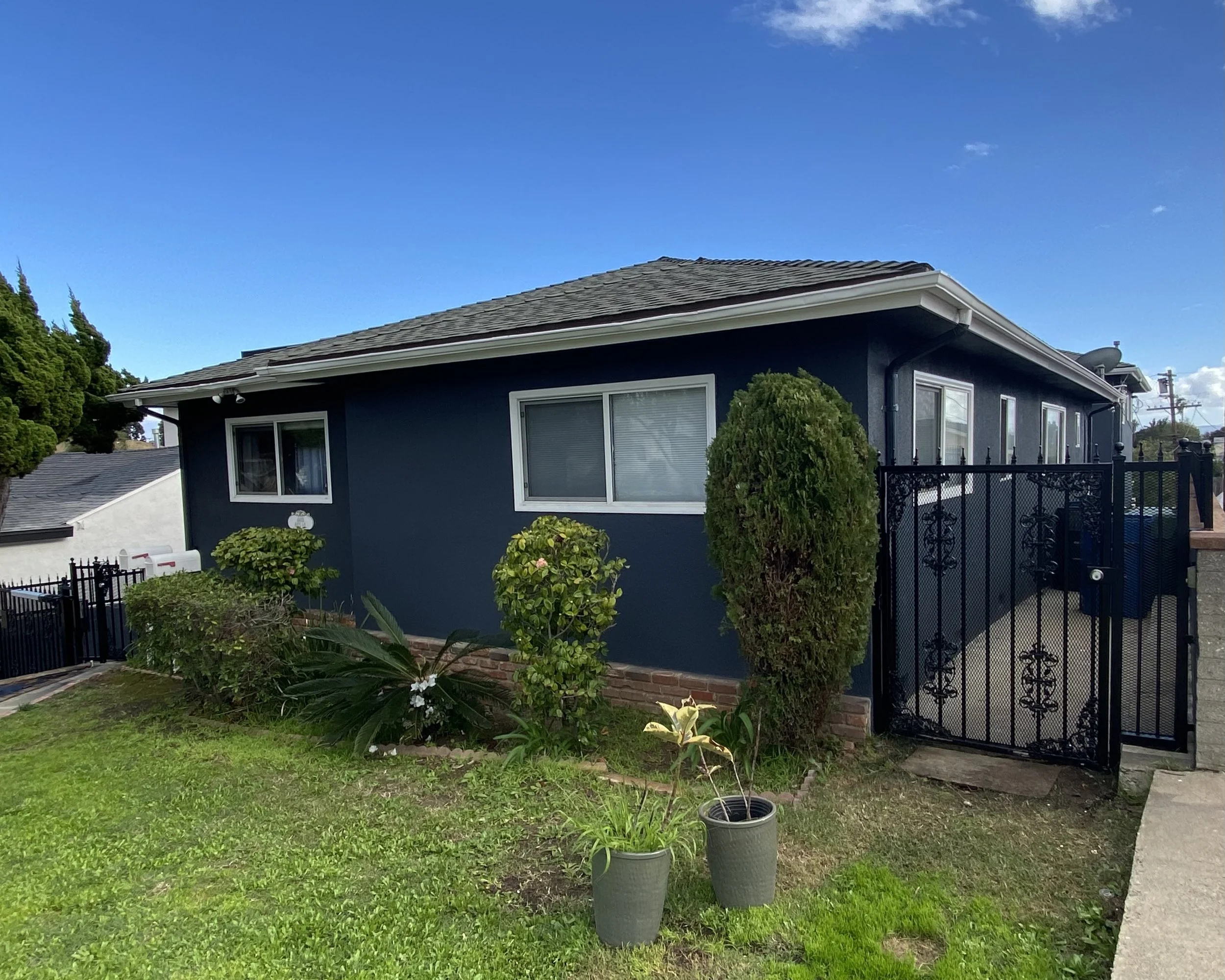 Dark blue single-story house with white trim, landscaped front yard, potted plants, and a decorative black metal gate under a bright blue sky