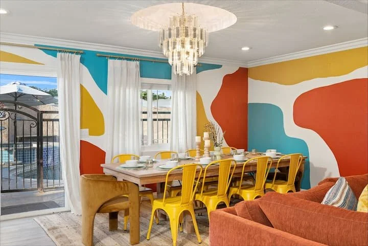 Colorful modern dining room with abstract mural walls, yellow metal chairs, crystal chandelier, and poolside patio doors