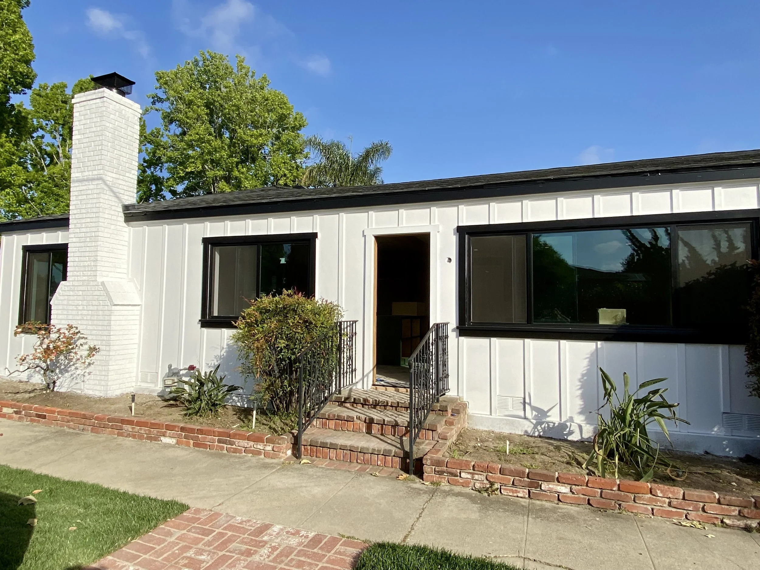 Sunny exterior of a renovated white mid-century home with black-trimmed windows, brick chimney, front steps, and landscaped yard