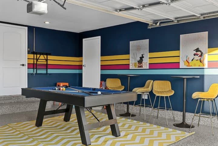Colorful retro game room with a pool table, yellow bar stools, striped walls, artwork, and a garage door