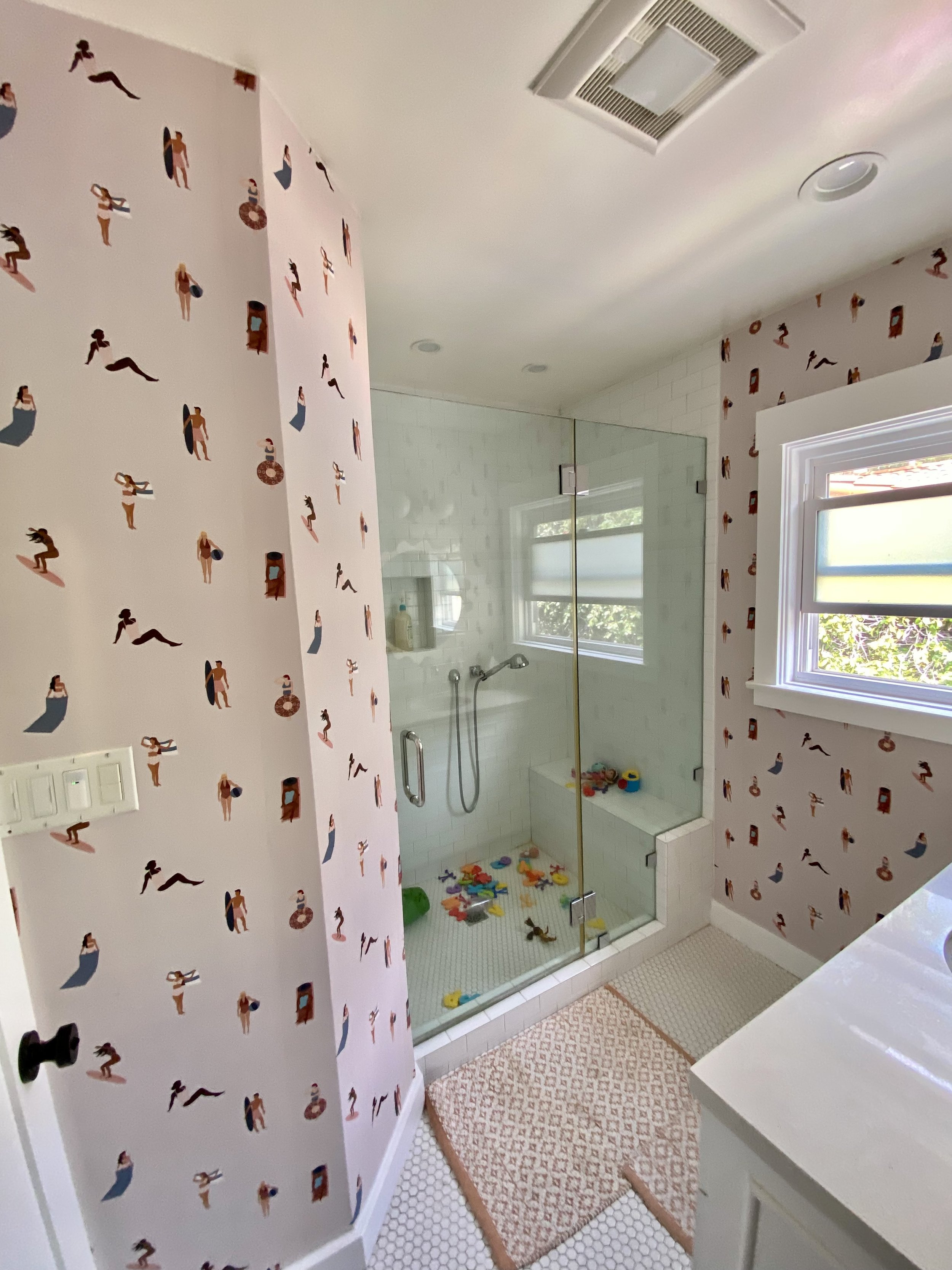 Bright children's bathroom with pink patterned wallpaper, glass shower enclosure, colorful bath toys, window, and patterned floor mats.