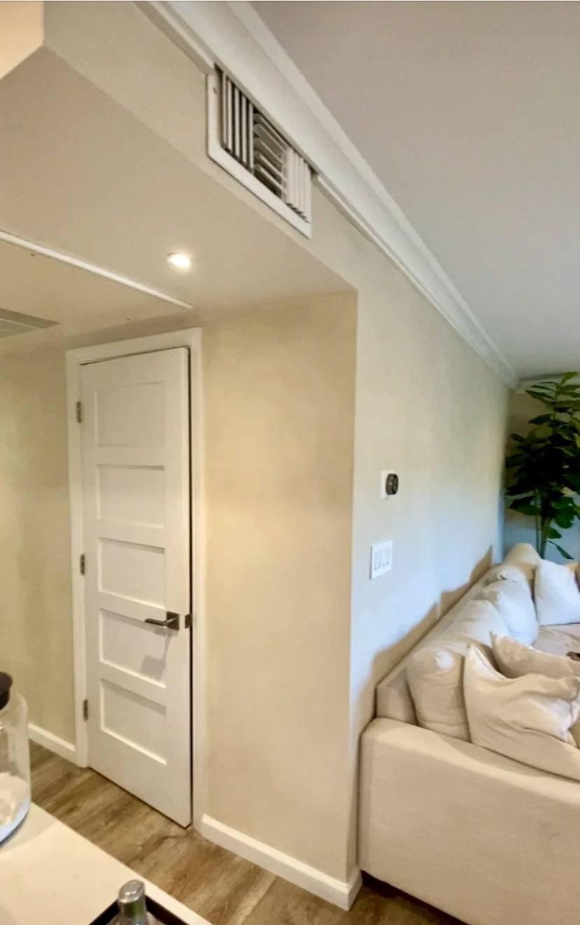 Bright modern living room with a white paneled door, wall vent, beige sectional sofa, recessed lighting, and indoor plant