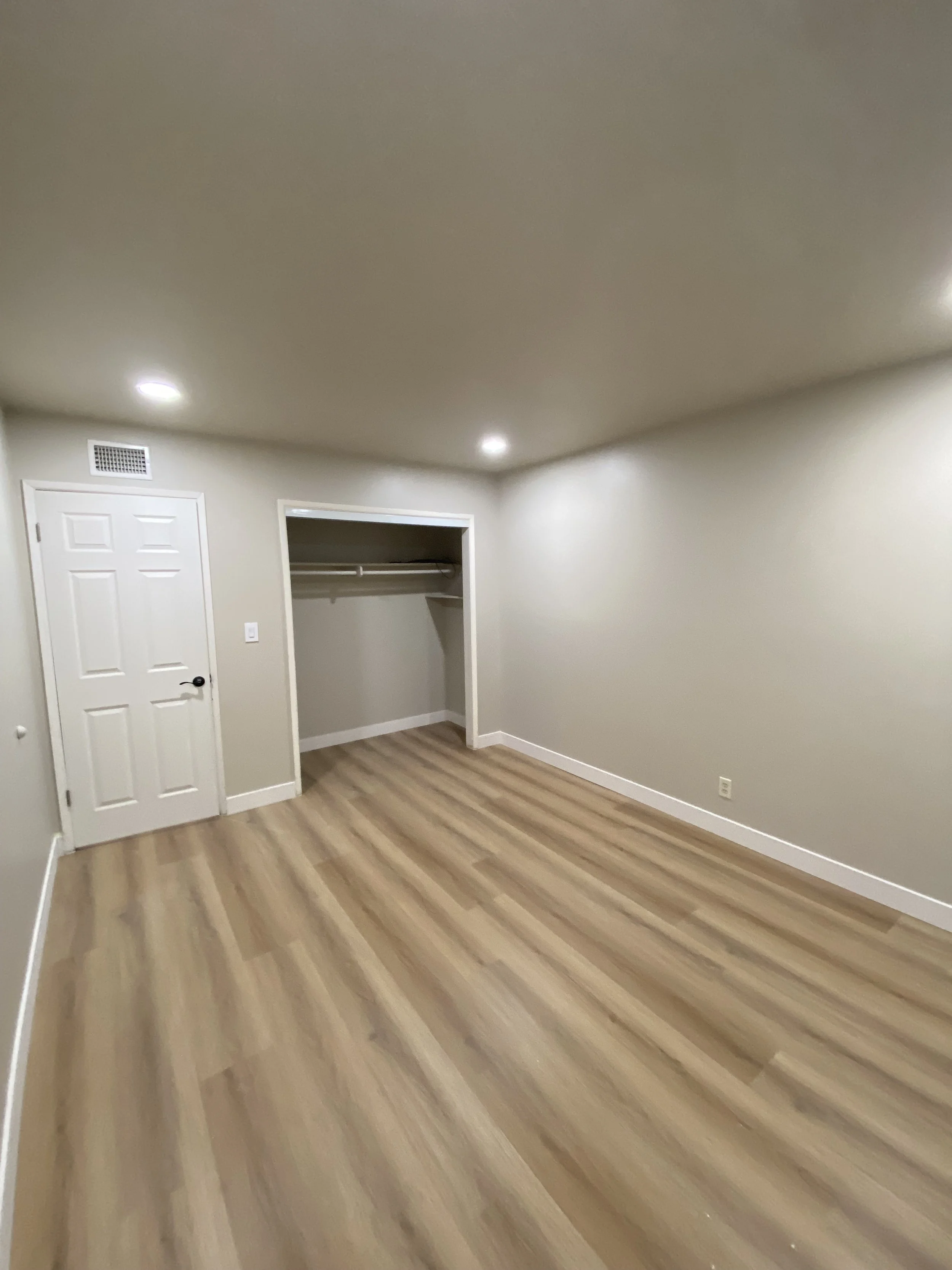 Empty beige room with wood-look flooring, recessed ceiling lights, a white door, and an open closet.