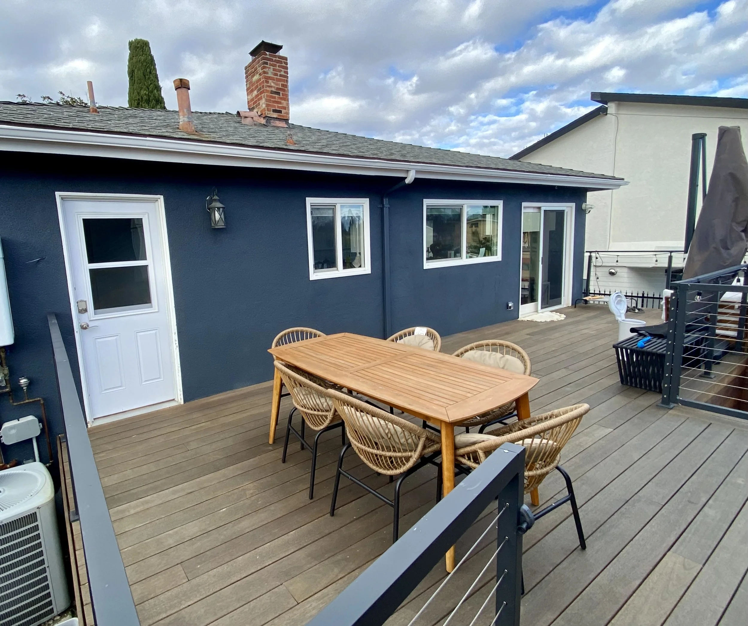 Outdoor rooftop deck with a wooden dining table, wicker chairs, dark blue house, brick chimney, grill, and cloudy sky
