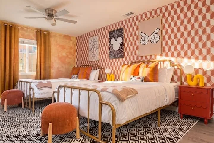 Retro-inspired bedroom with two beds, geometric wallpaper and rug, orange accents, and colorful wall art