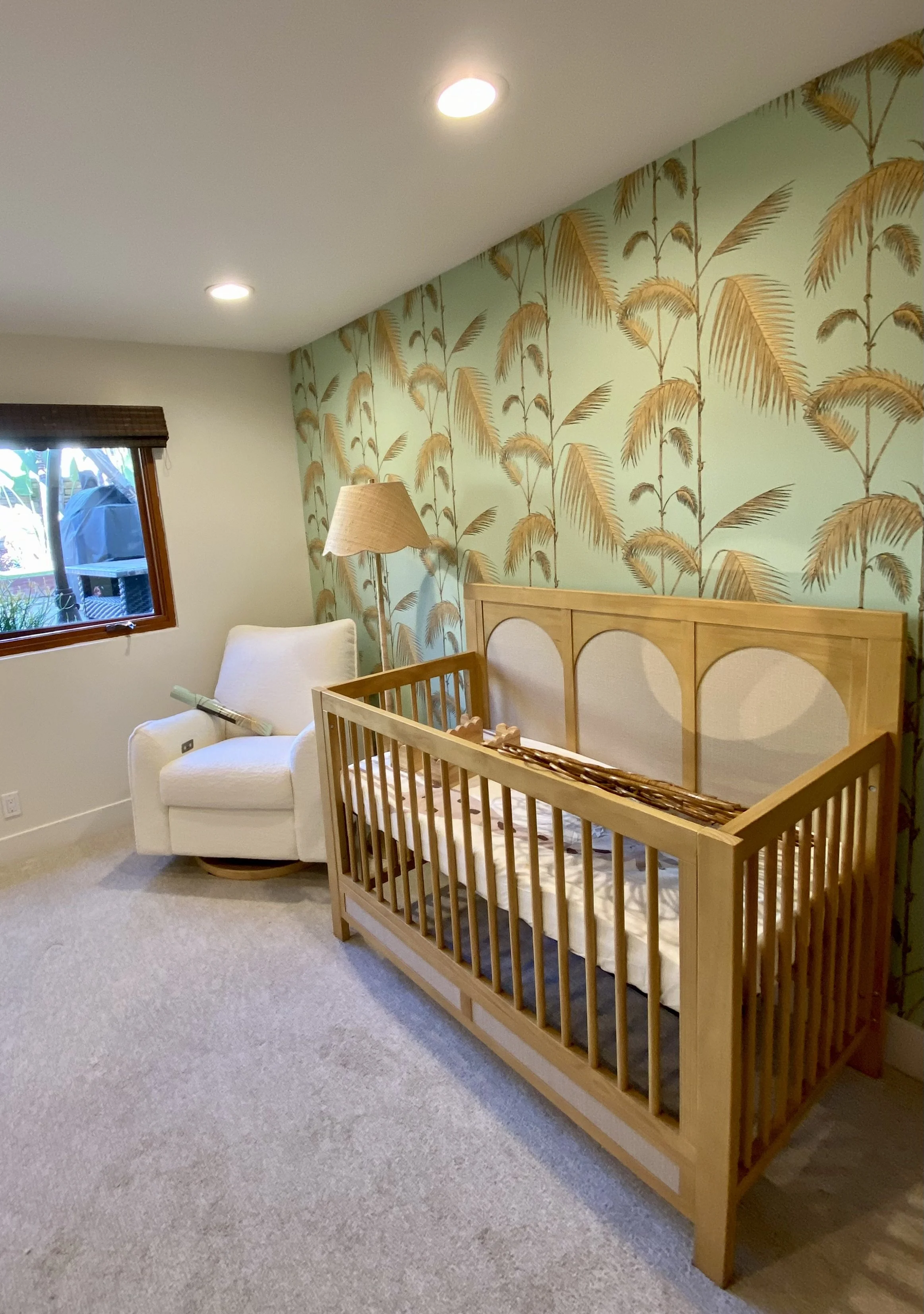 Nursery with a wooden crib, white armchair, floor lamp, window, and tropical palm tree wallpaper
