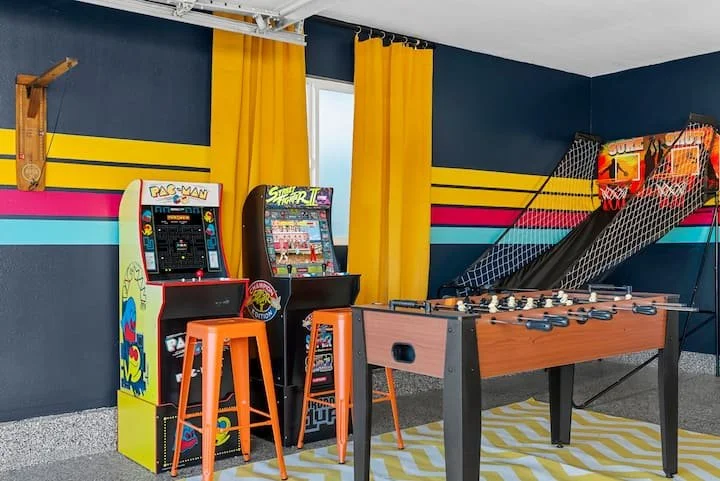 Retro game room with Pac-Man and Street Fighter arcade cabinets, orange stools, foosball table, and basketball hoops