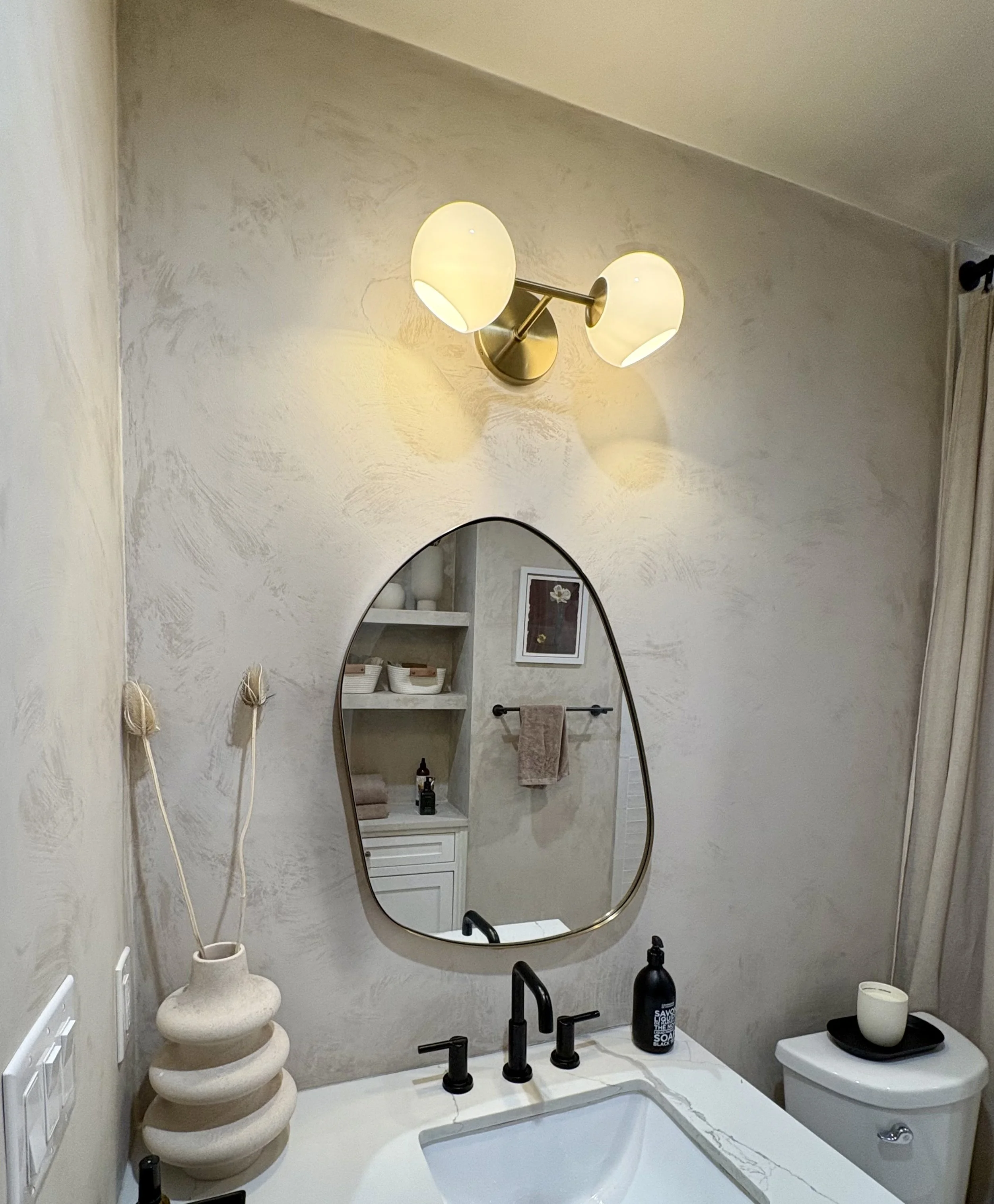 Modern bathroom vanity with black fixtures, illuminated oval mirror, globe wall light, decorative vase, and toilet