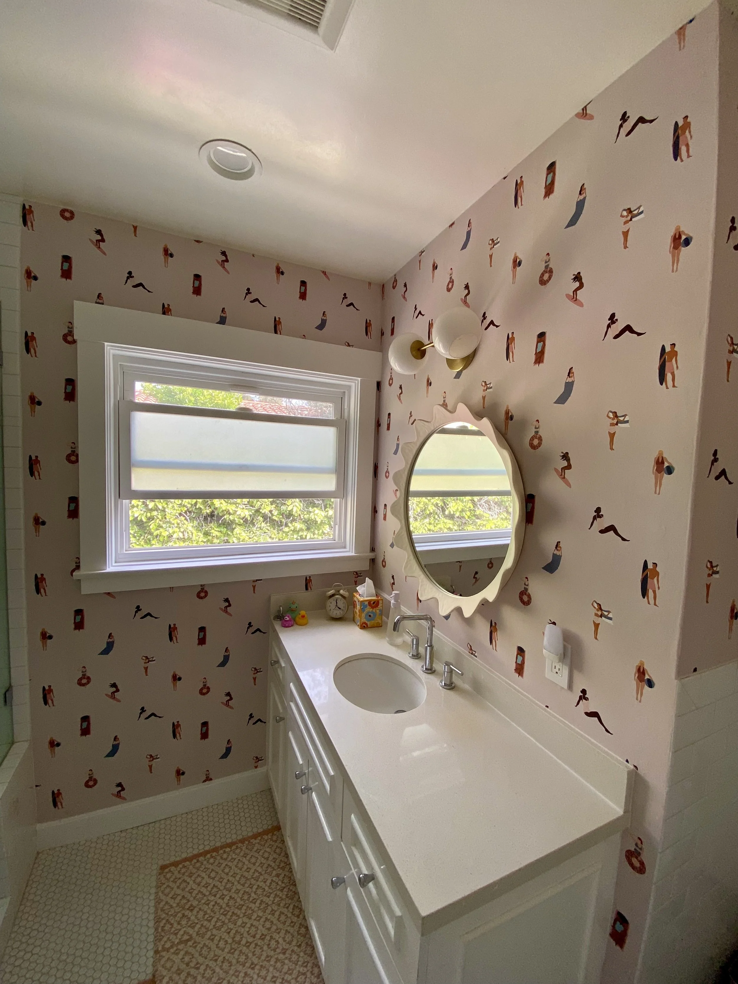 Bright bathroom with patterned wallpaper, white vanity and sink, scalloped mirror, window, bathtub, and decorative light fixture.