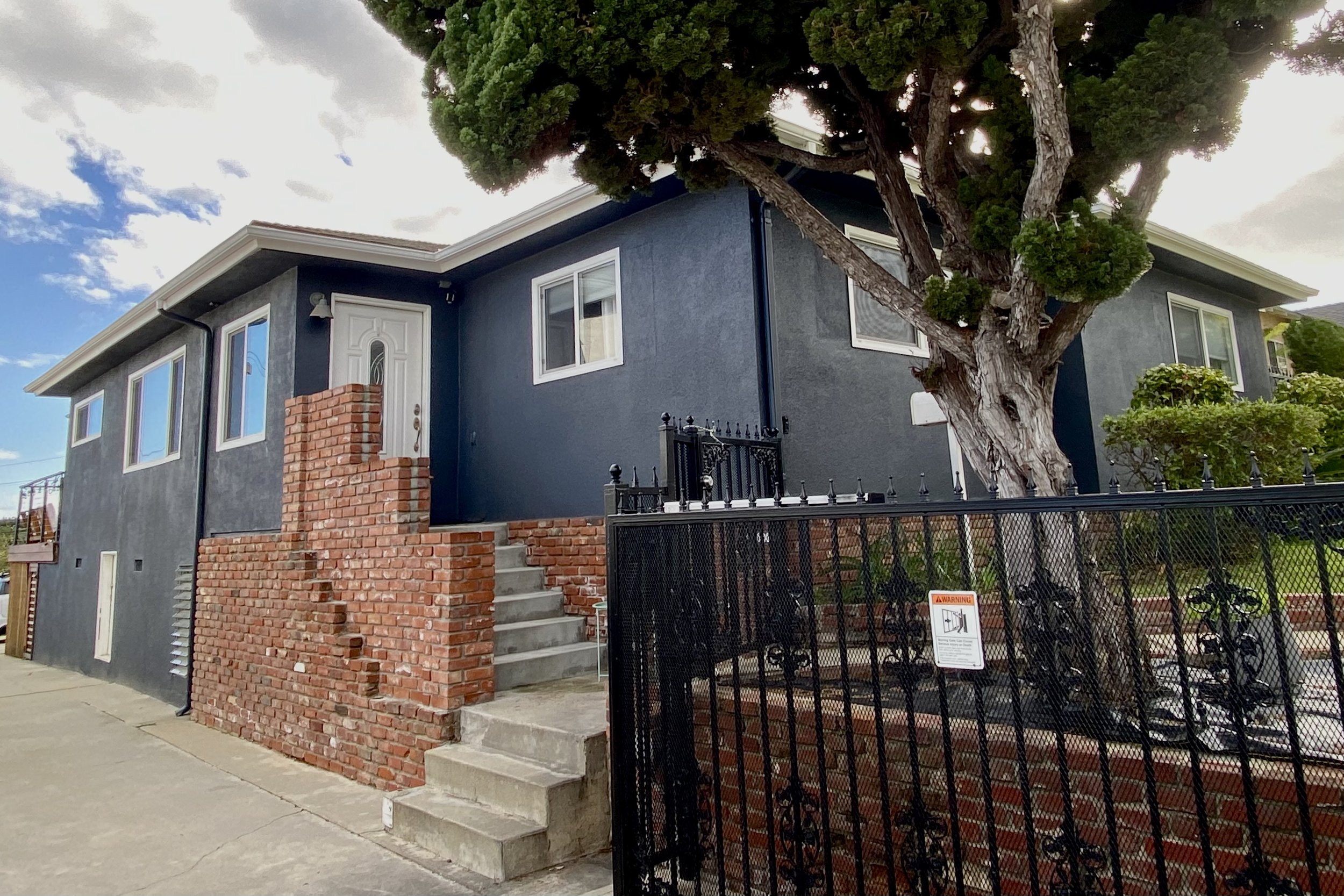 Dark gray stucco house with brick entry steps, white-trimmed windows, manicured trees, and a black metal fence.