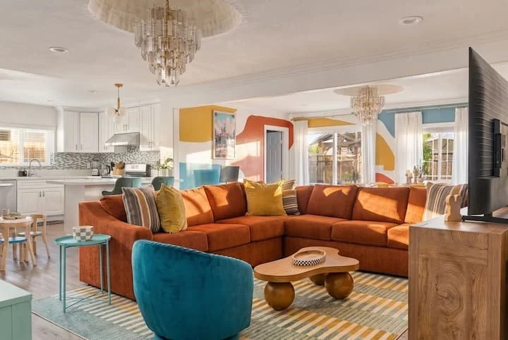 Bright colorful open-plan living room with orange sectional sofa, turquoise chair, patterned rug, coffee table, kitchen, and crystal chandeliers