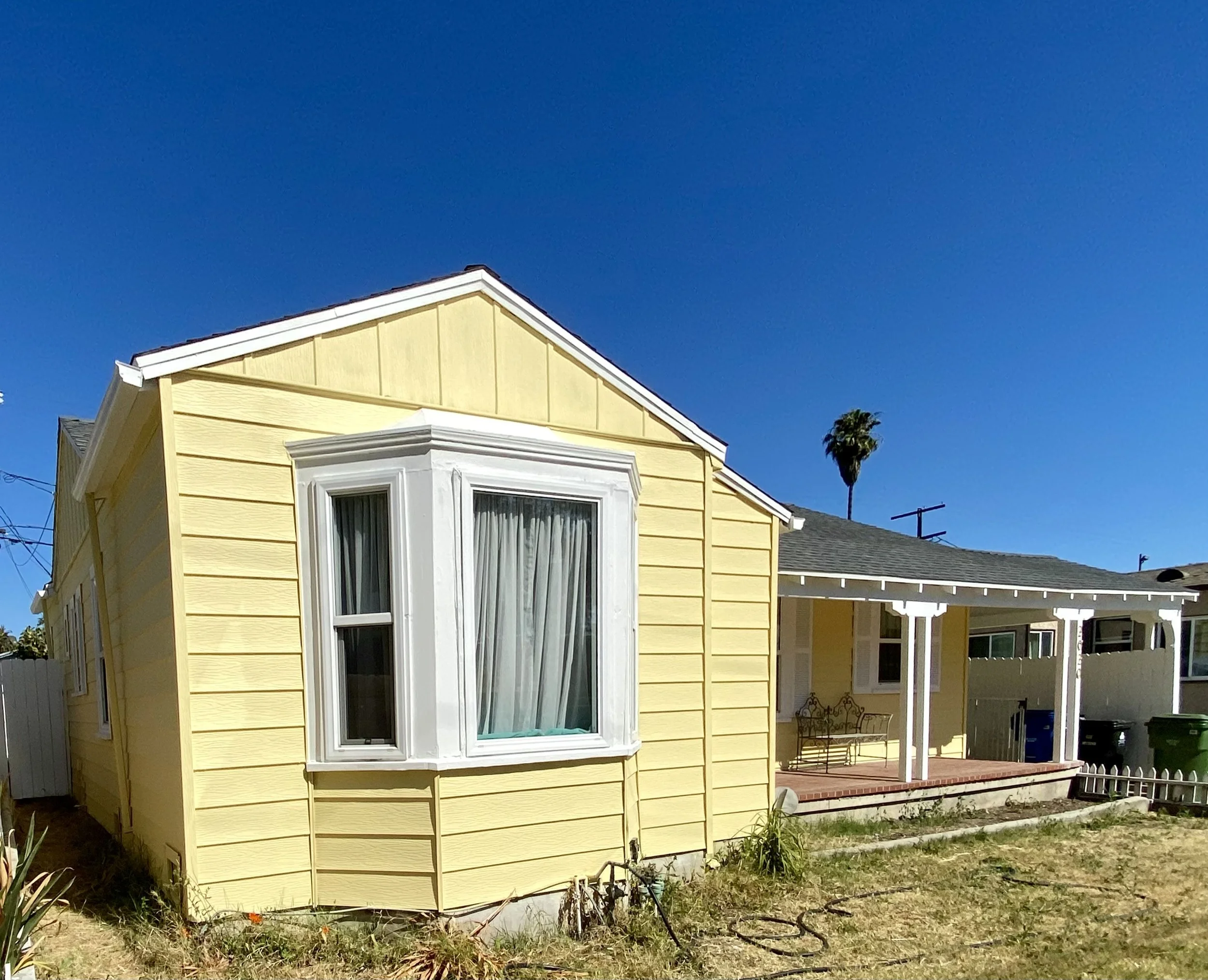 Yellow single-story house with white trim, bay window, covered patio, and palm tree under a clear blue sky