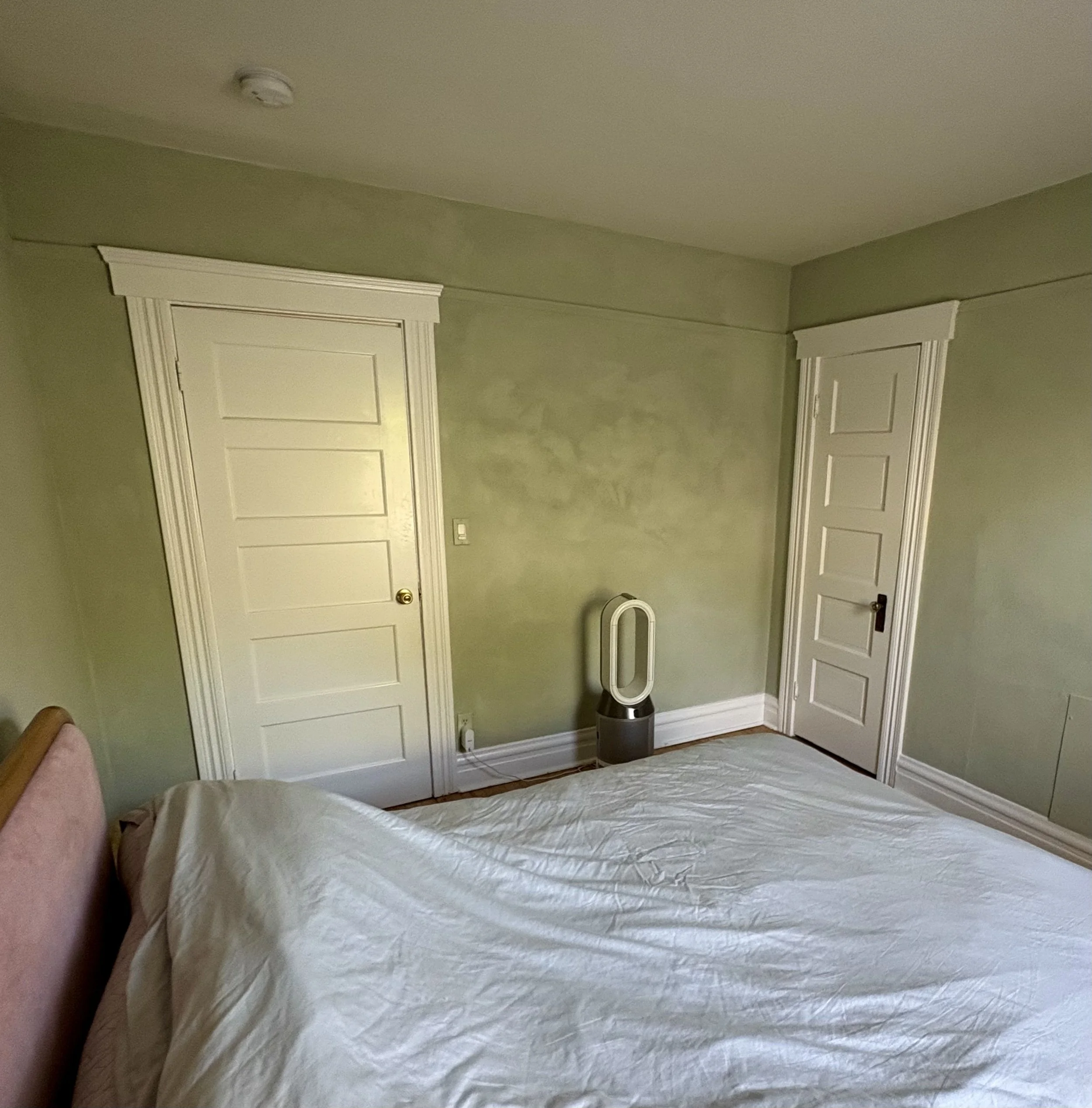 Bedroom with green walls, two white paneled doors, a white bed, and a bladeless tower fan.