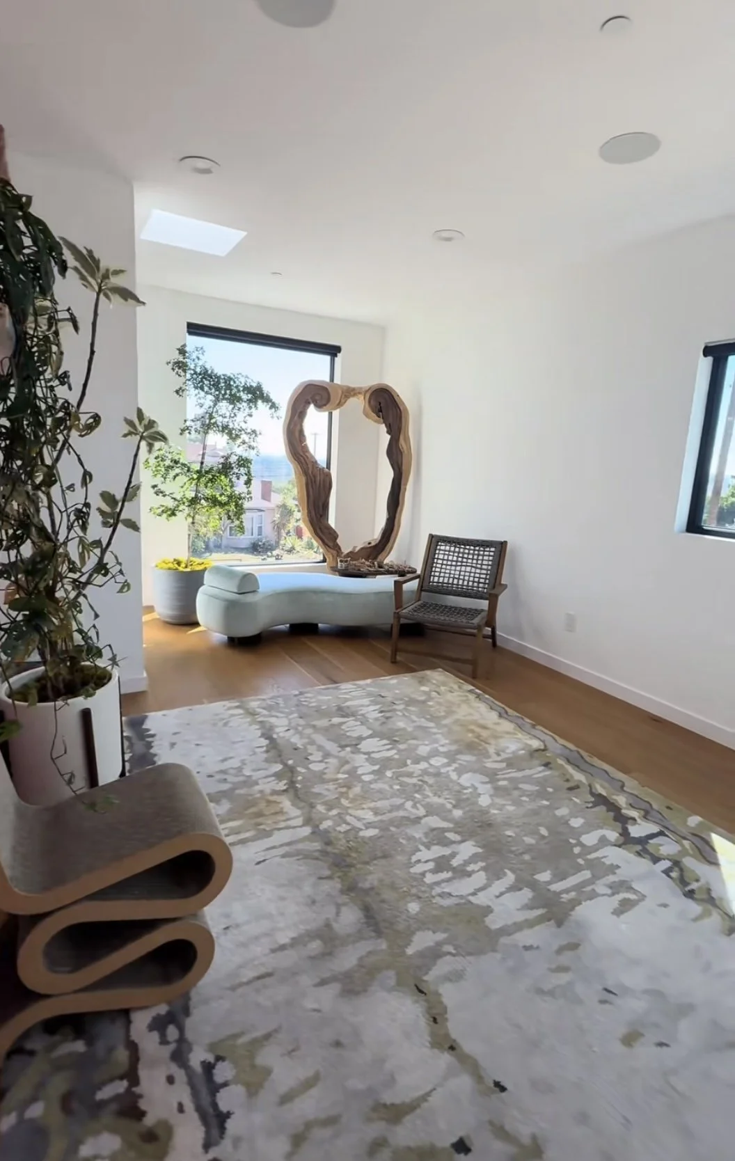 Bright modern living room with a large abstract rug, curved lounge chair, indoor plants, sculptural wooden mirror, and chaise lounge by the windows
