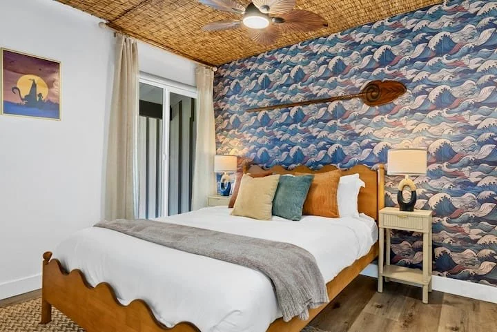 Colorful coastal-themed bedroom with wave-patterned wallpaper, wooden bed, white bedding, ceiling fan, and bedside table
