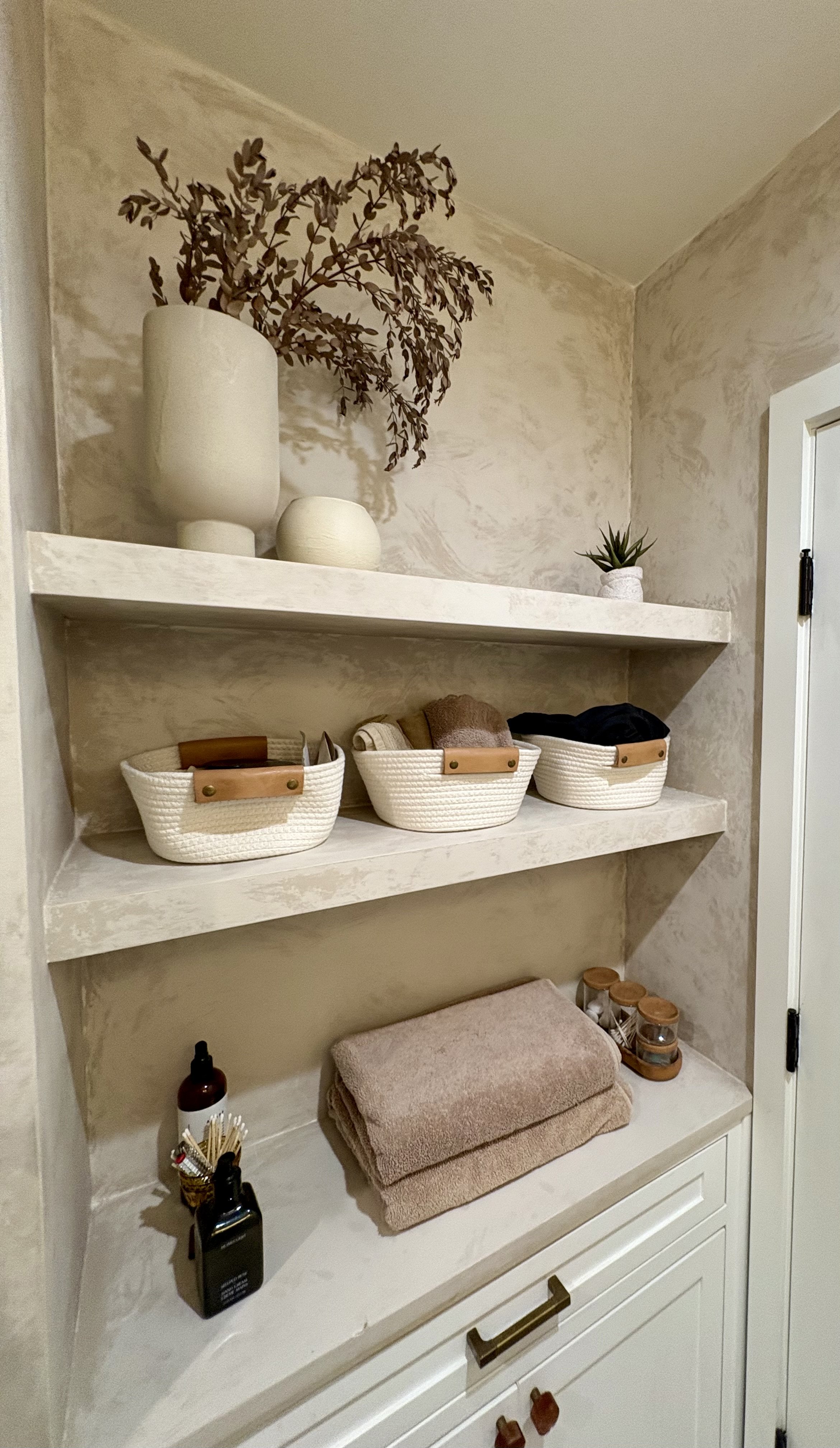 Built-in bathroom shelving with woven baskets, folded towels, toiletries, decorative plants, and a hanging leafy plant.