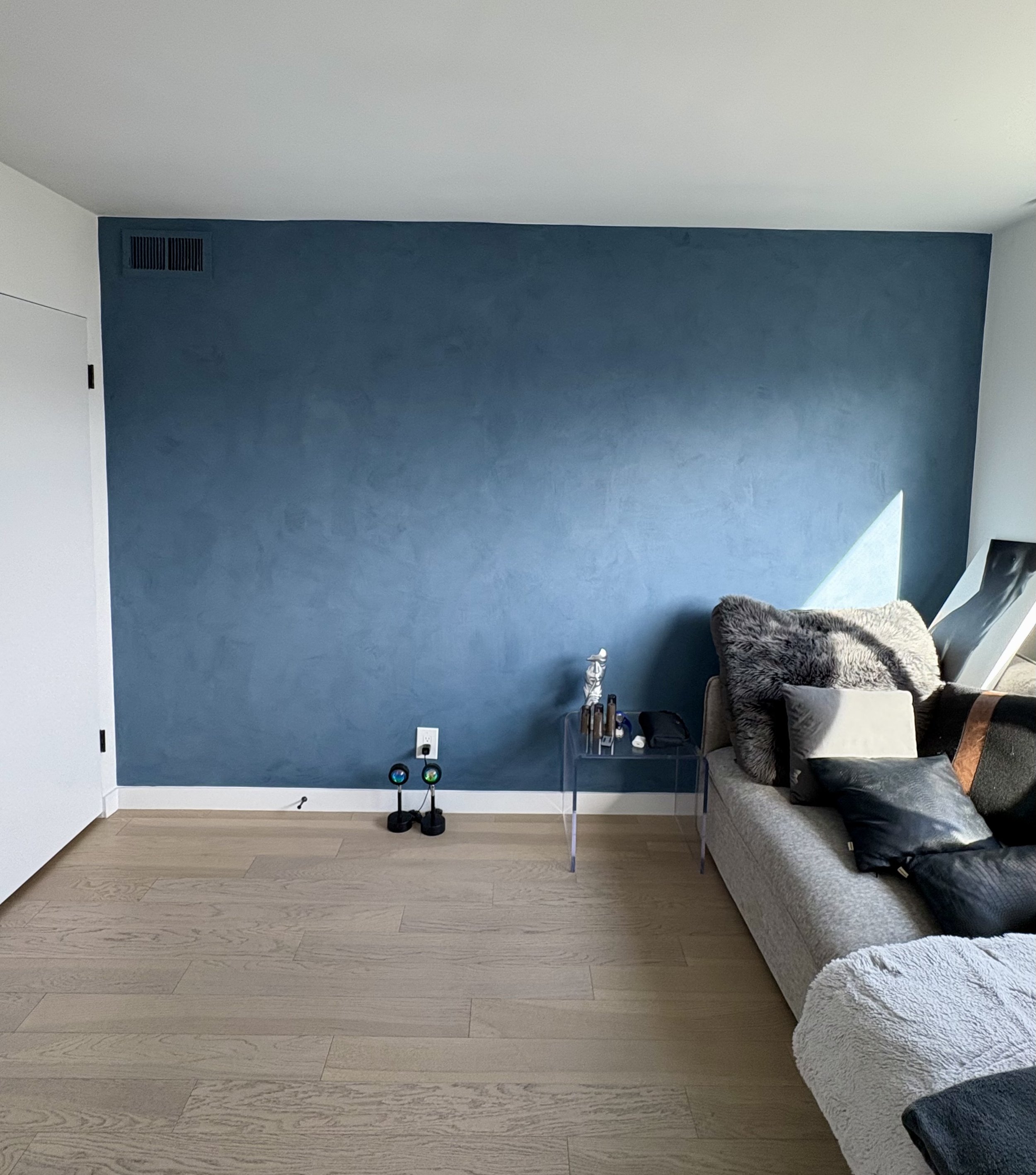 Modern living room with a dark blue accent wall, gray sectional sofa, glass side table, decorative lamps, and light wood flooring