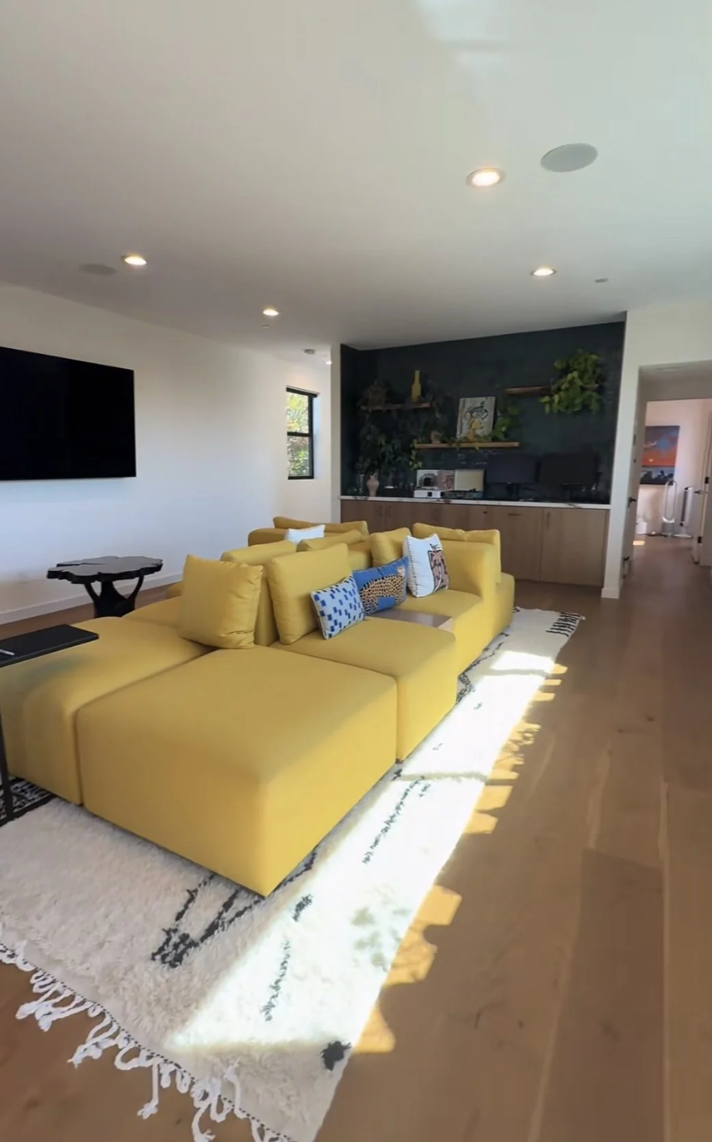 Bright modern living room with a large yellow sectional sofa, patterned rug, wall-mounted TV, and dark built-in entertainment center.