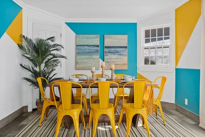 Bright dining room with yellow metal chairs, wooden table, tropical plant, ocean artwork, and turquoise geometric walls