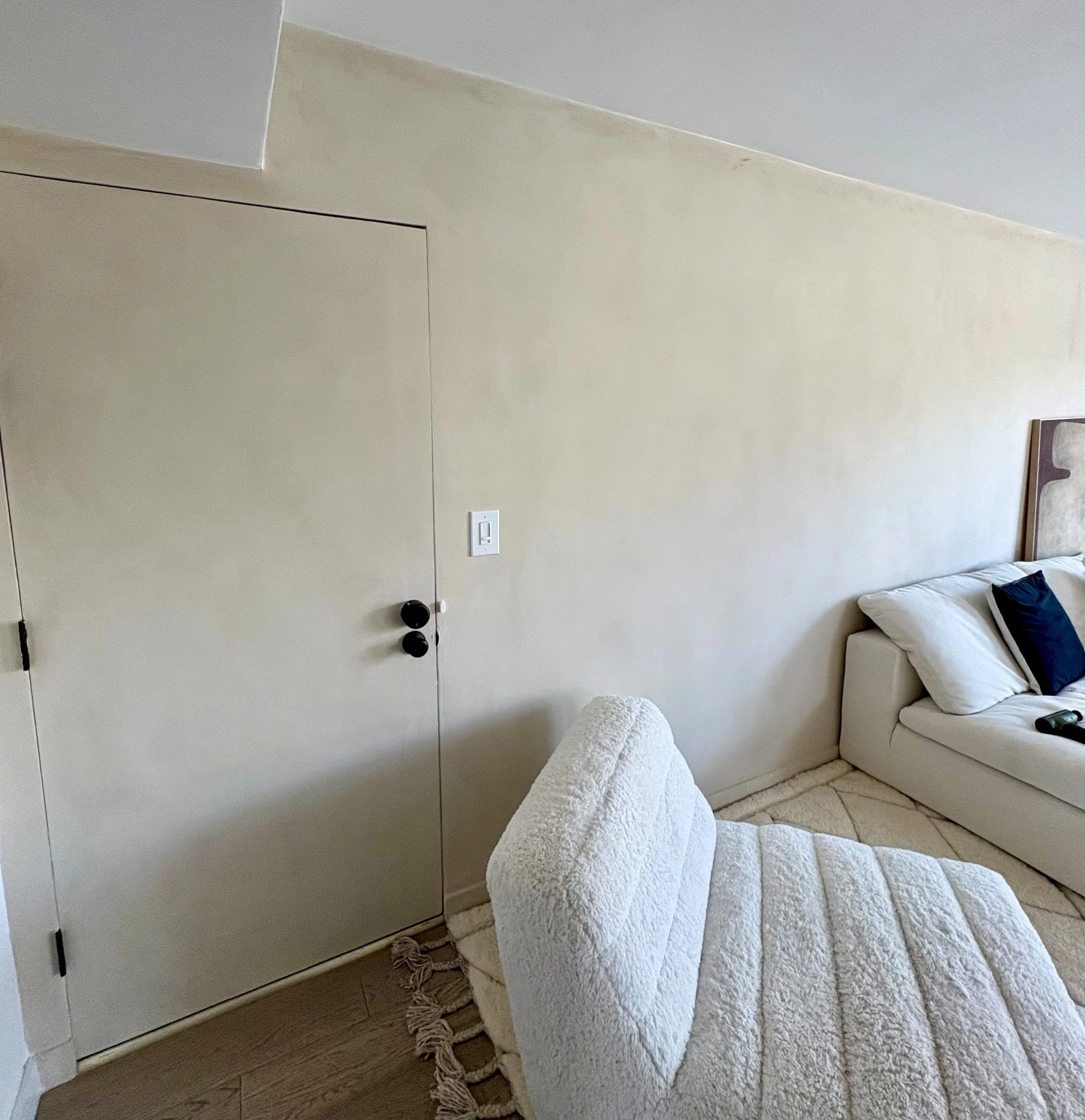 Bright modern bedroom with a white bed, textured blanket, pillows, upholstered bench, wall-mounted light switch, and closed double doors.