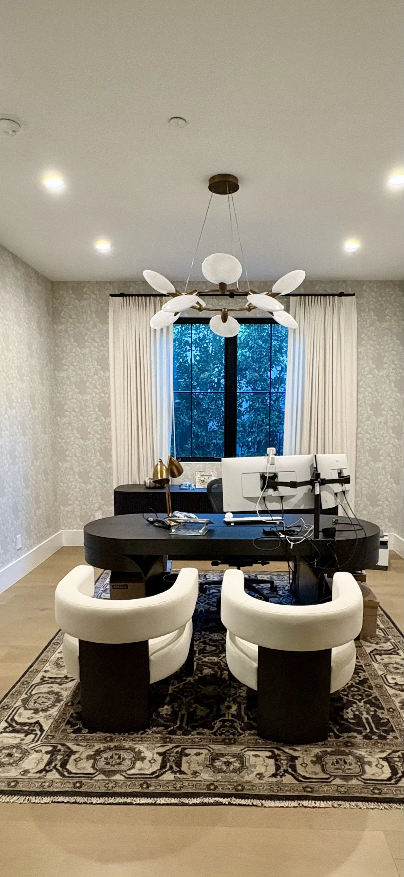 Modern home office with a black desk, computer monitors, white chairs, patterned rug, floral wallpaper, and sculptural ceiling light.
