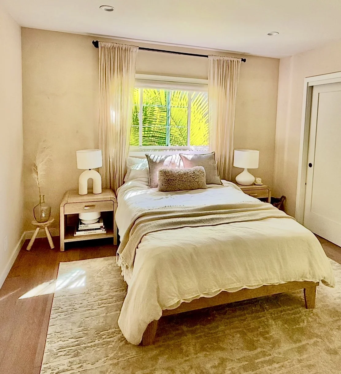 Bright, cozy bedroom with a neatly made bed, cream bedding, bedside tables and lamps, and a window overlooking tropical palm leaves.