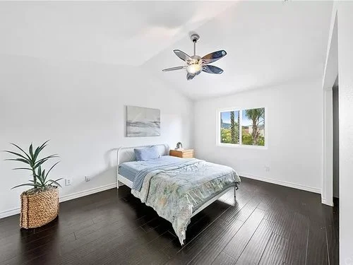 Bright modern bedroom with a neatly made bed, dark wood floors, ceiling fan, potted plant, and sunny window