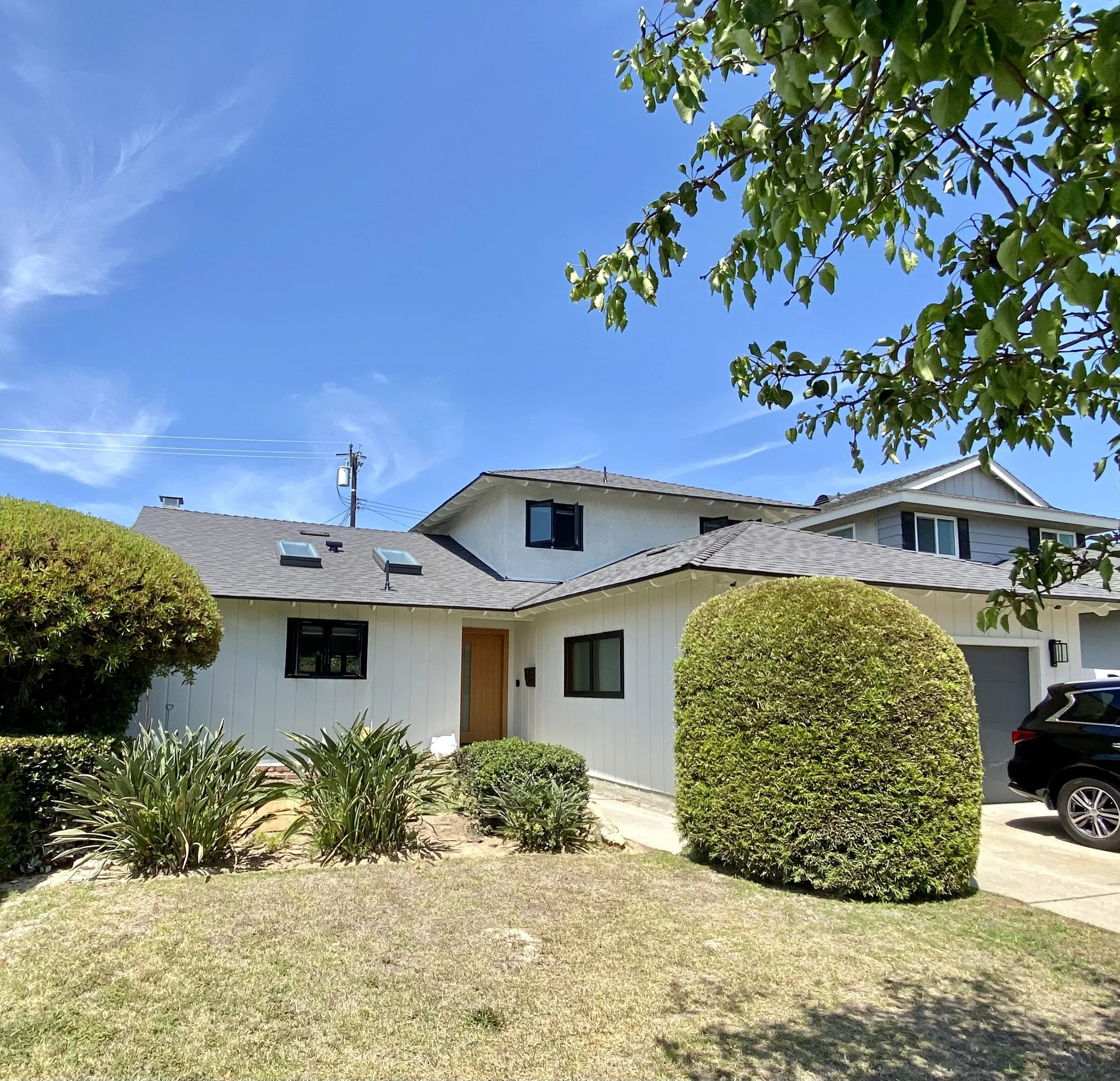 White suburban house with gray roof, manicured hedges, landscaped front yard, and parked black SUV under a bright blue sky