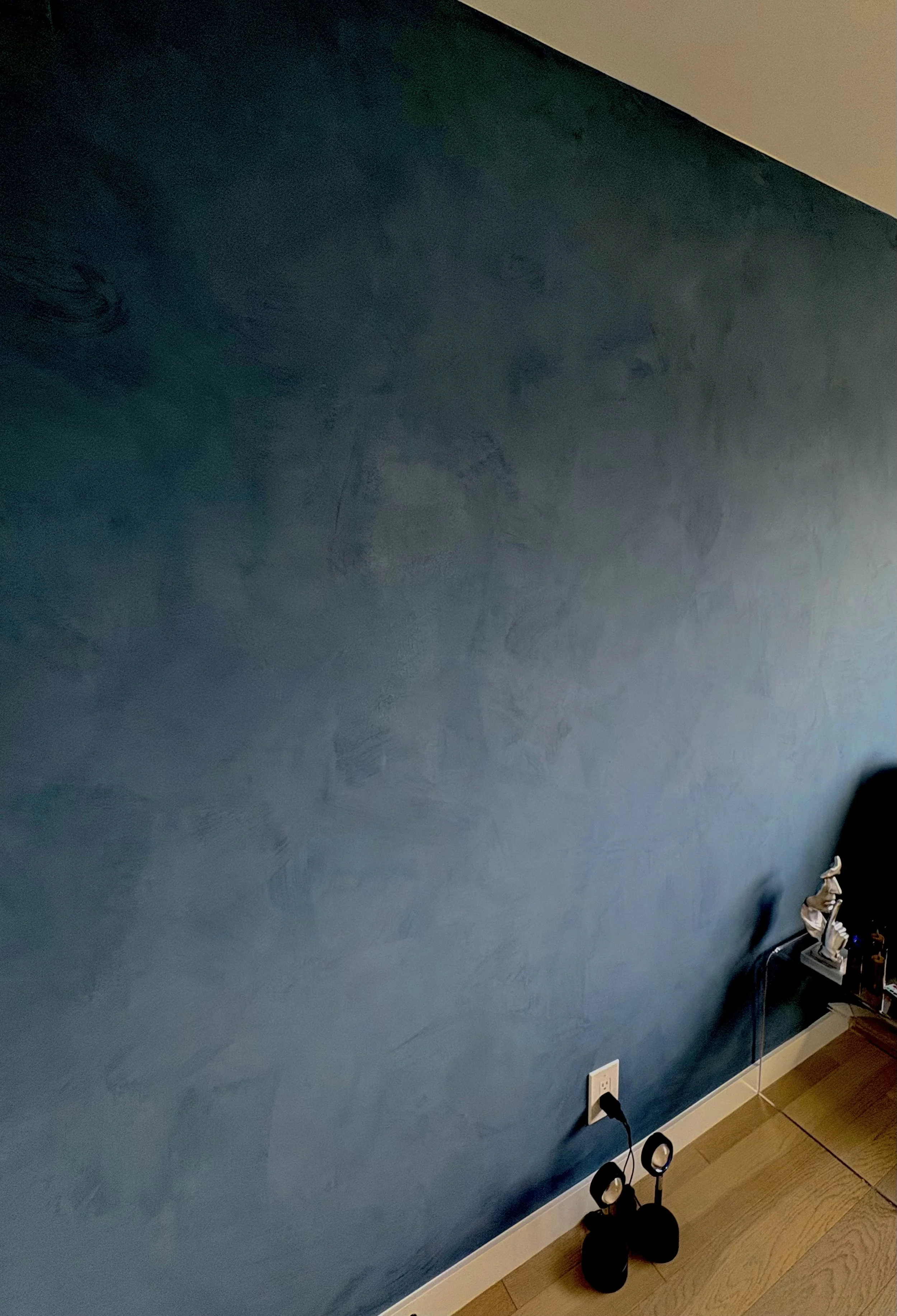 Blue-painted interior wall with white baseboard, electrical outlet, hardwood floor, black floor lamp, and small figurine.