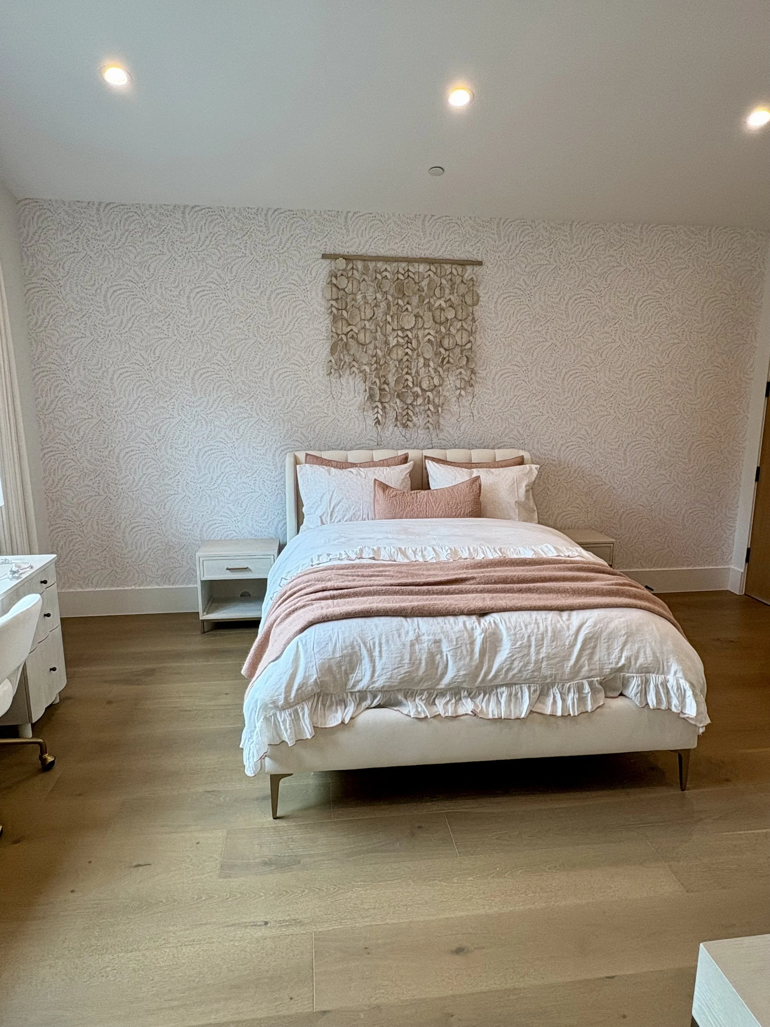 Elegant bedroom with a white upholstered bed, pink and white bedding, patterned wallpaper, shell wall hanging, nightstands, and recessed ceiling lights.