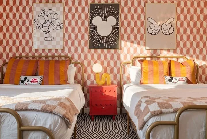 Retro twin bedroom with colorful striped bedding, Mickey Mouse wall art, patterned wallpaper, red nightstand, and geometric rug
