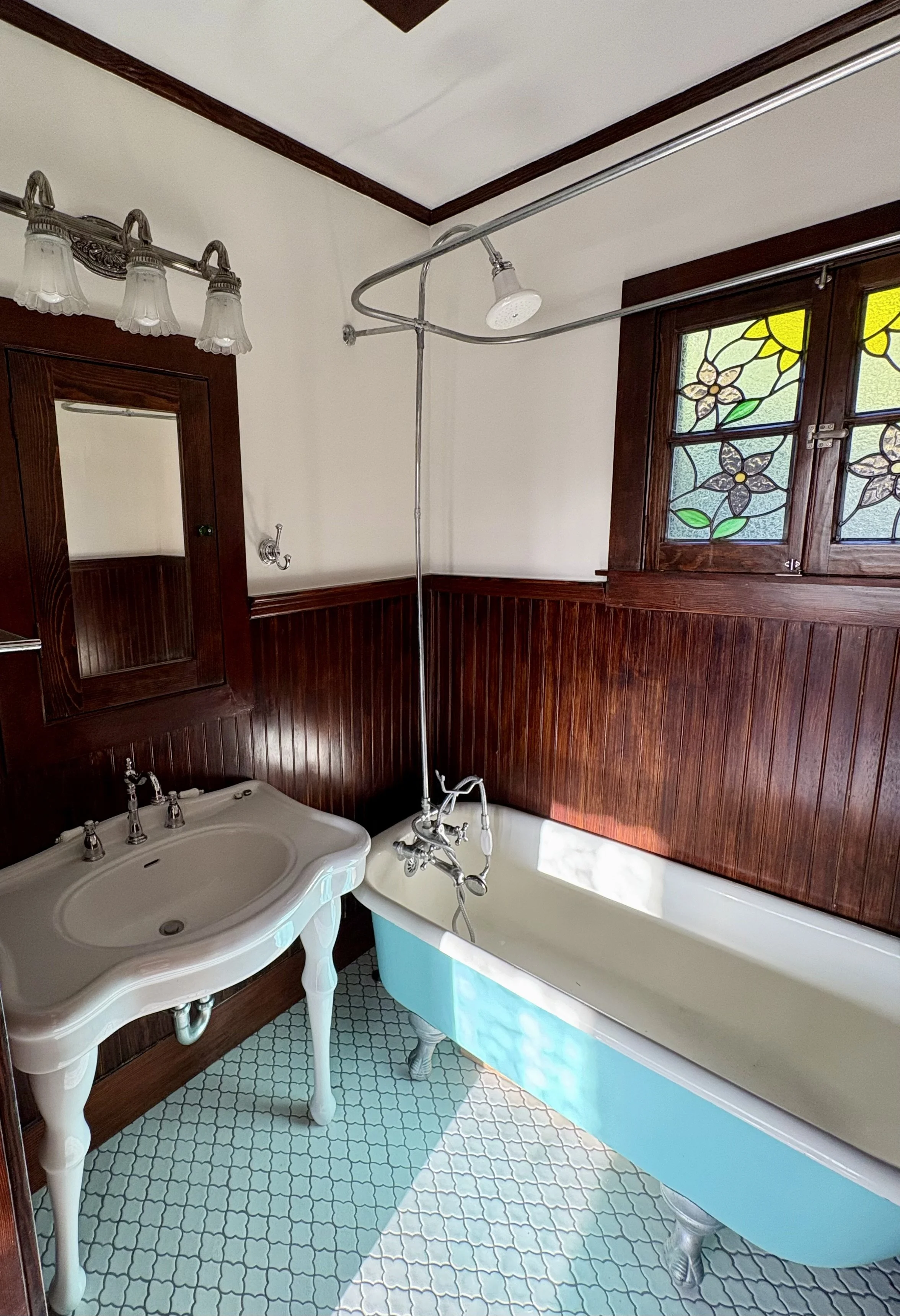 Vintage bathroom with a clawfoot bathtub, pedestal sink, dark wood wainscoting, stained-glass windows, and exposed shower plumbing.