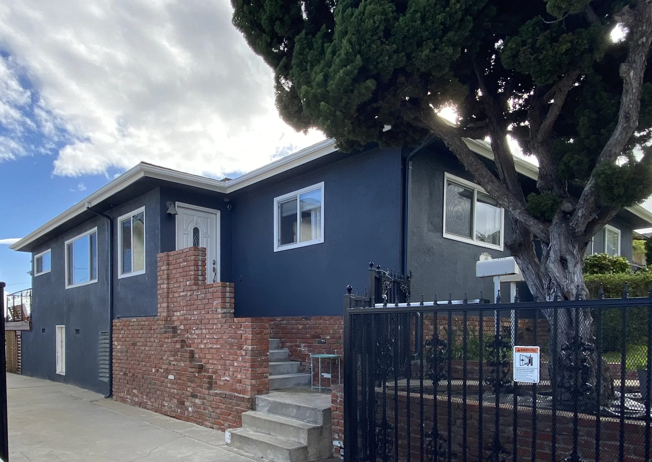 Dark gray stucco house with white trim, brick entry wall, fenced front yard, and large evergreen tree.