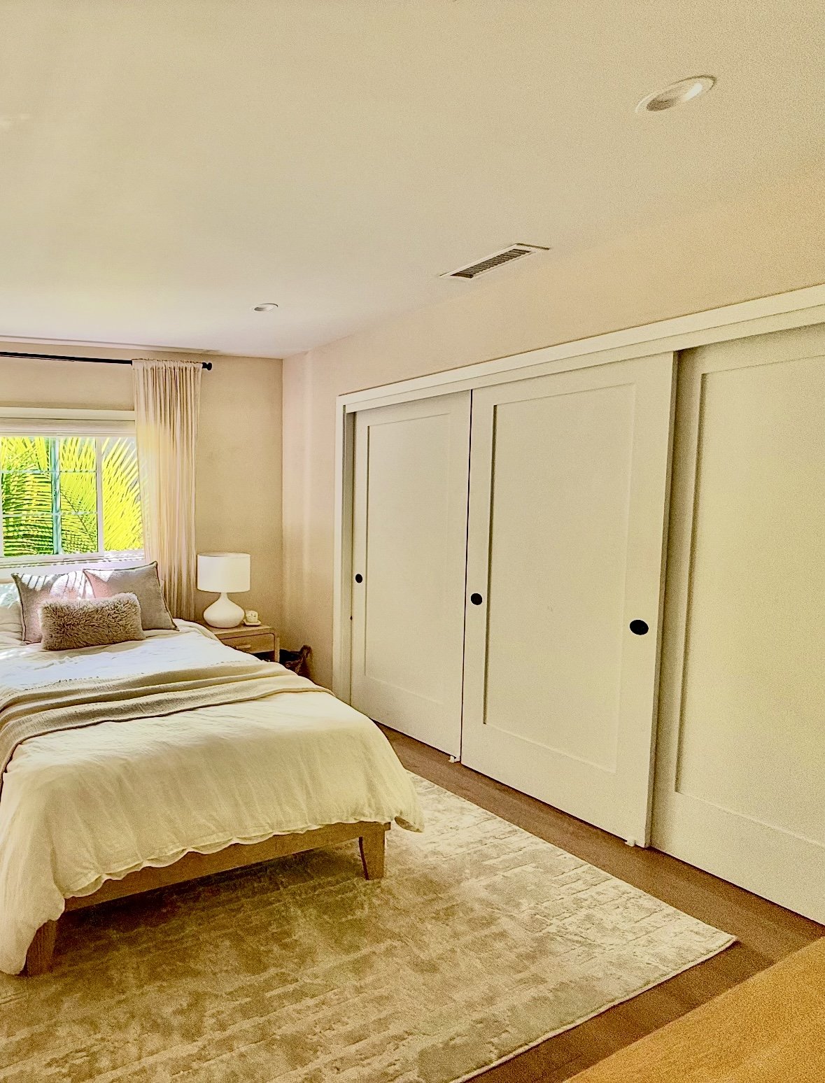 Bright neutral-toned bedroom with a neatly made bed, bedside lamp, window, area rug, and large white sliding-door closet.