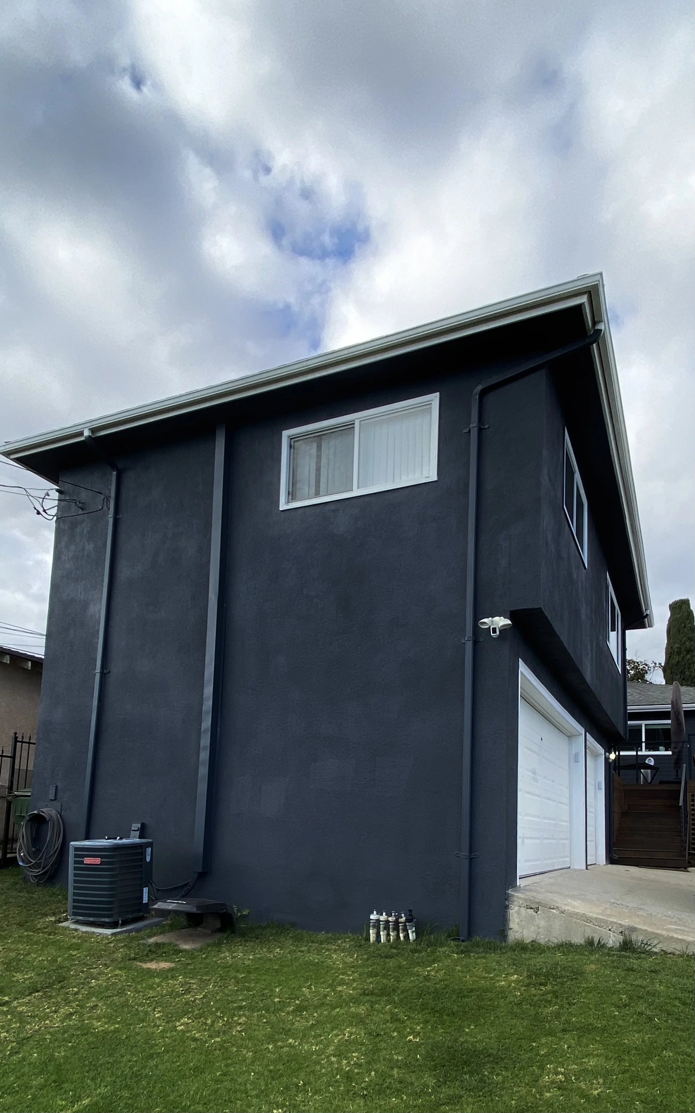 Dark gray stucco house exterior with white-trimmed windows, garage doors, rooftop gutters, and an outdoor air-conditioning unit under a cloudy sky.