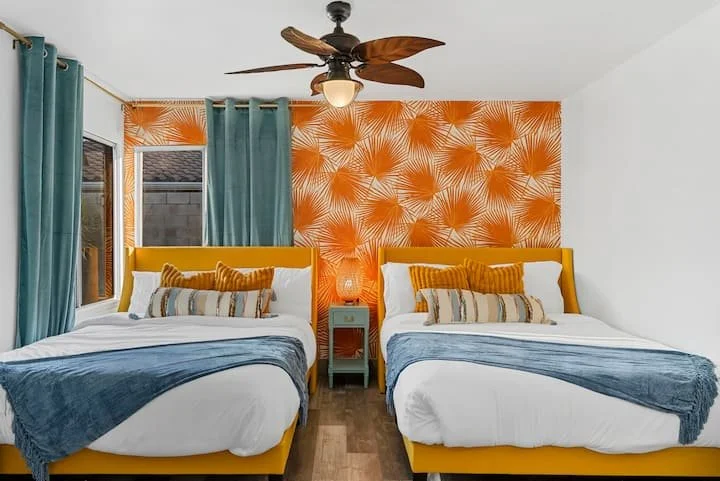 Bright bedroom with two mustard-yellow beds, blue curtains and blankets, orange palm leaf wallpaper, and a ceiling fan
