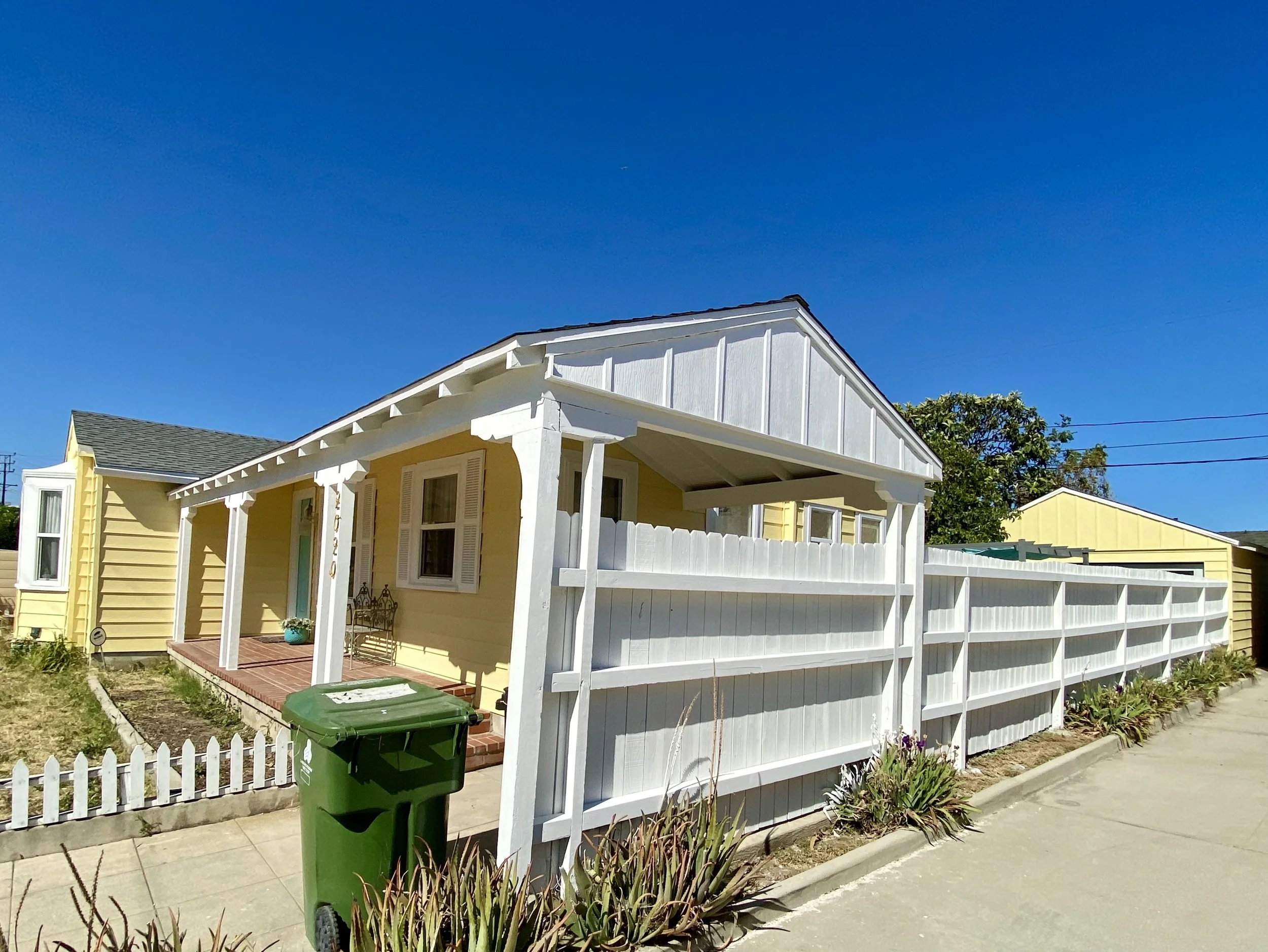 Sunny residential street with a yellow cottage, white trim and porch, picket fence, and green trash bin under a clear blue sky