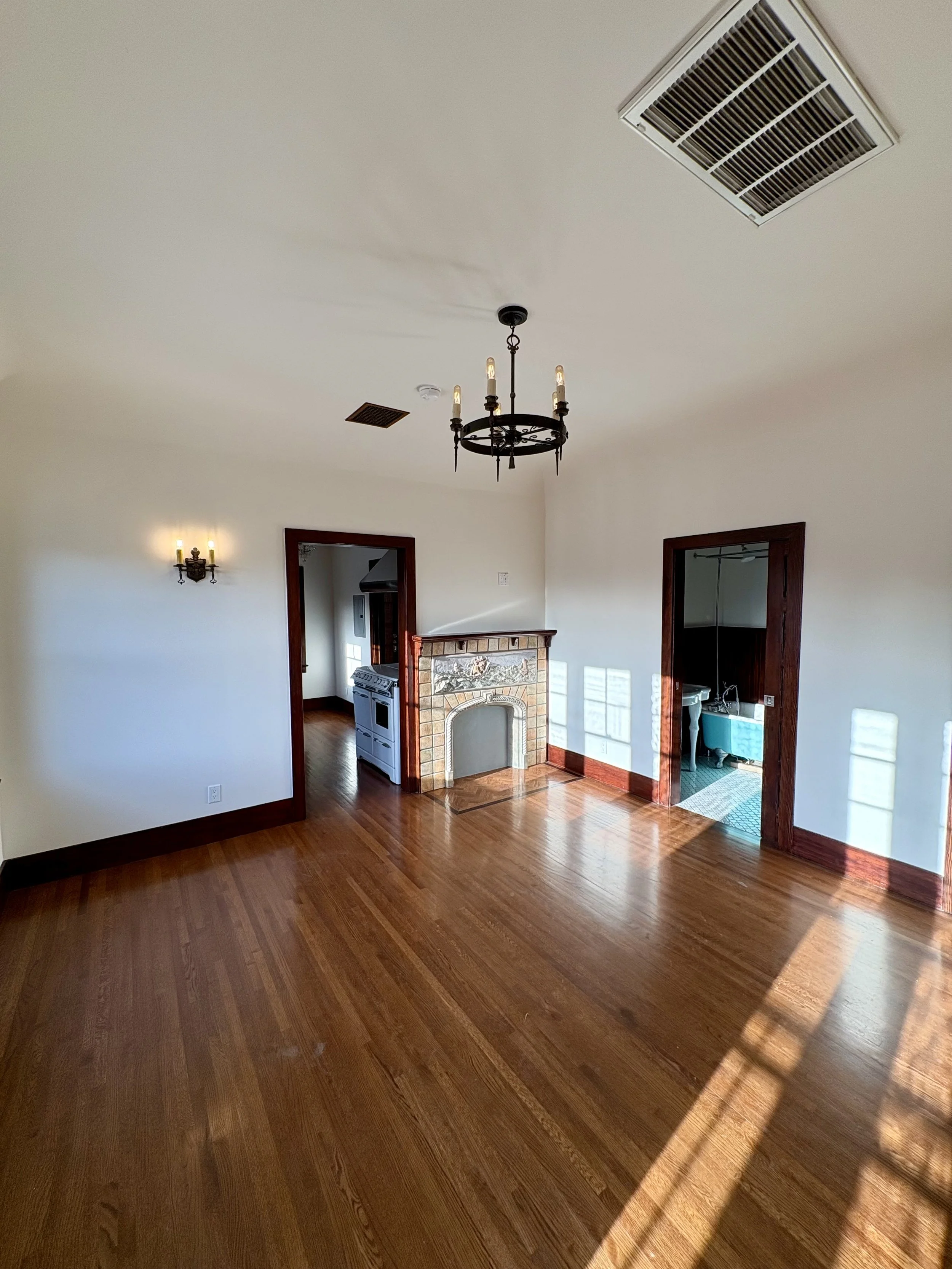 Sunlit vintage living room with hardwood floors, ornate chandelier, tiled fireplace, and adjoining kitchen and bathroom doors