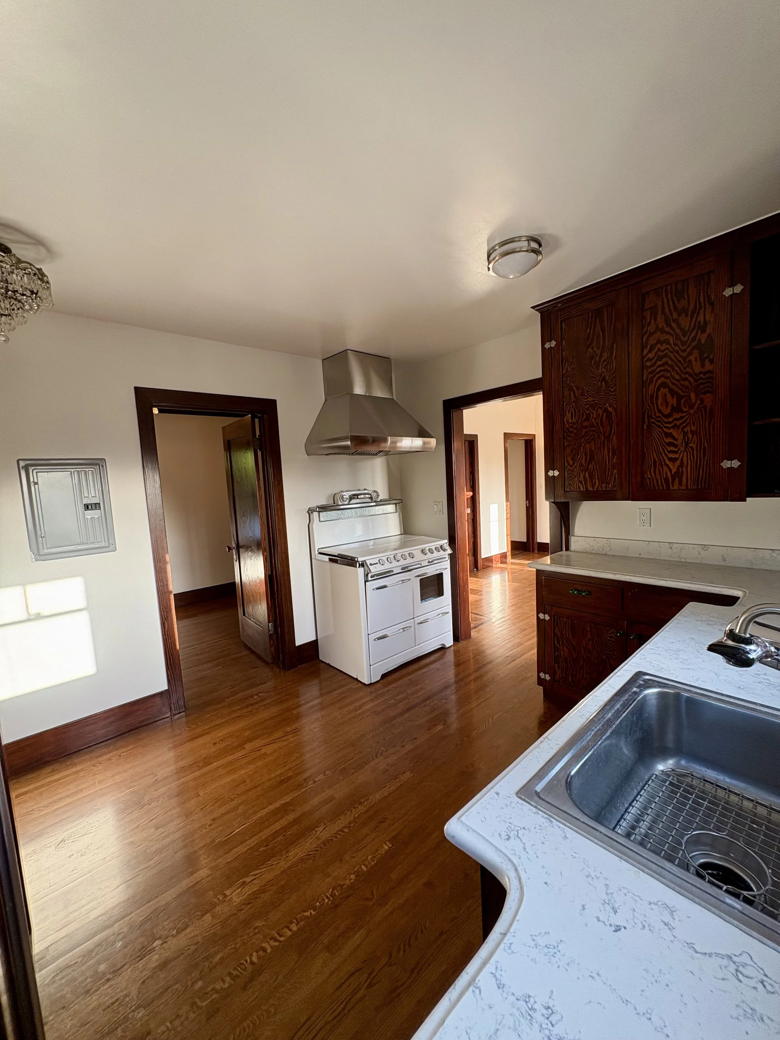 Spacious vintage kitchen with dark wood cabinets, white stove and range hood, marble-look countertops, farmhouse sink, and hardwood floors.