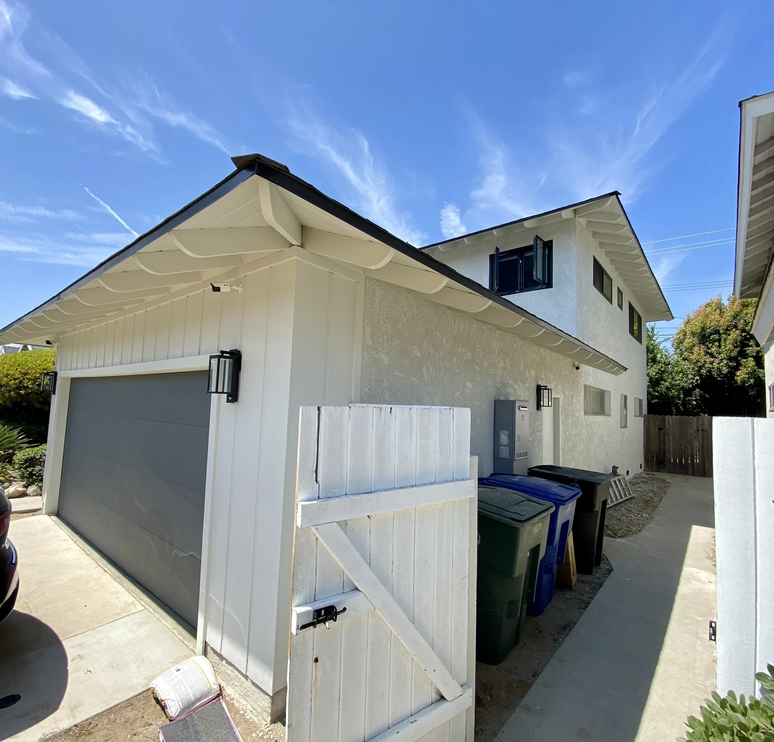 White residential garage and house exterior with a gray garage door, fenced side yard, trash bins, and blue sky