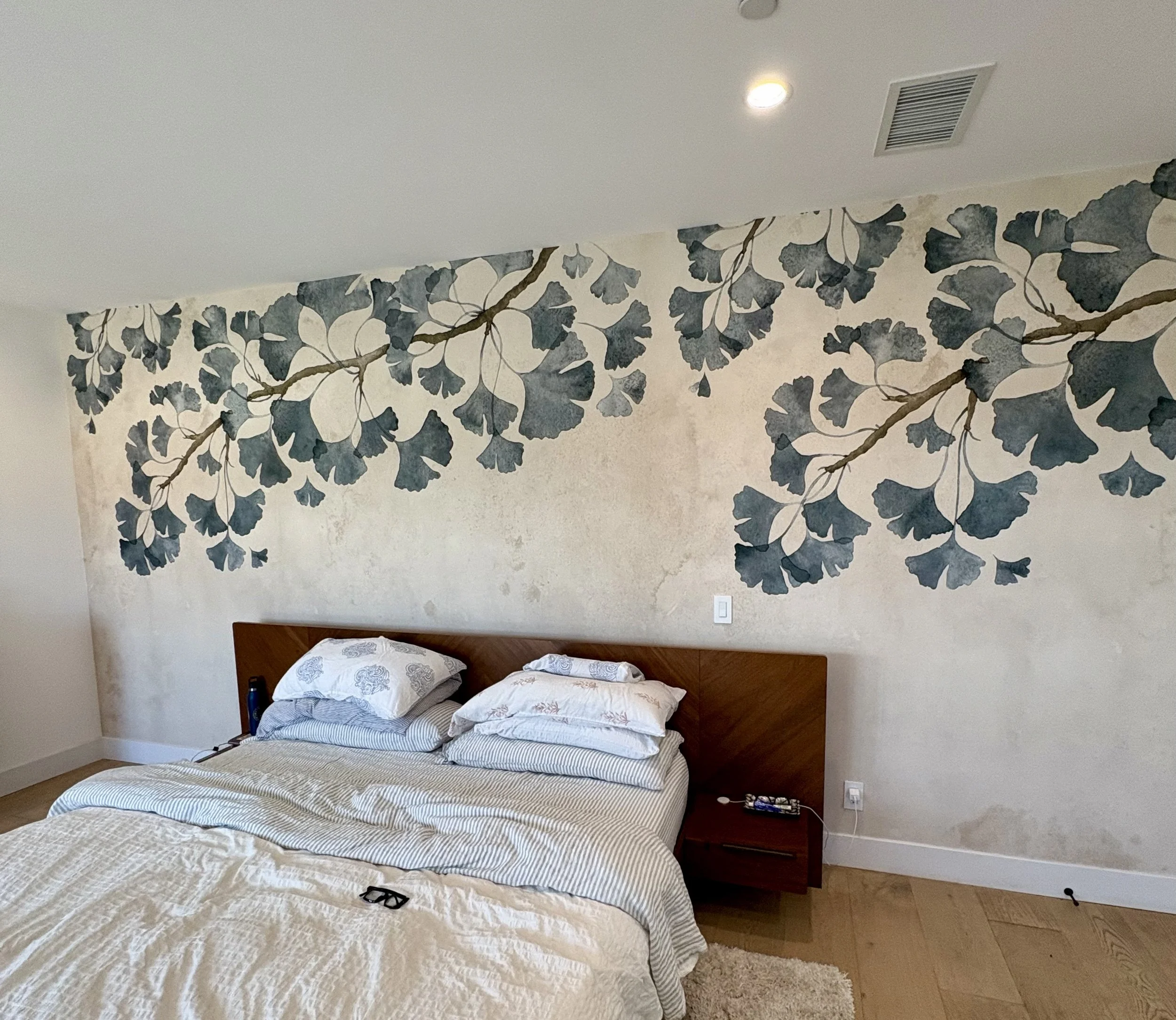 Bedroom with a wooden bed, white striped bedding, and blue botanical wallpaper featuring ginkgo branches.