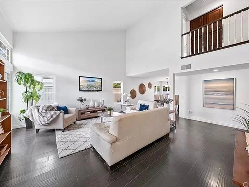 Bright modern living room with white sofas, dark hardwood floors, wall-mounted TV, artwork, and an upstairs balcony railing