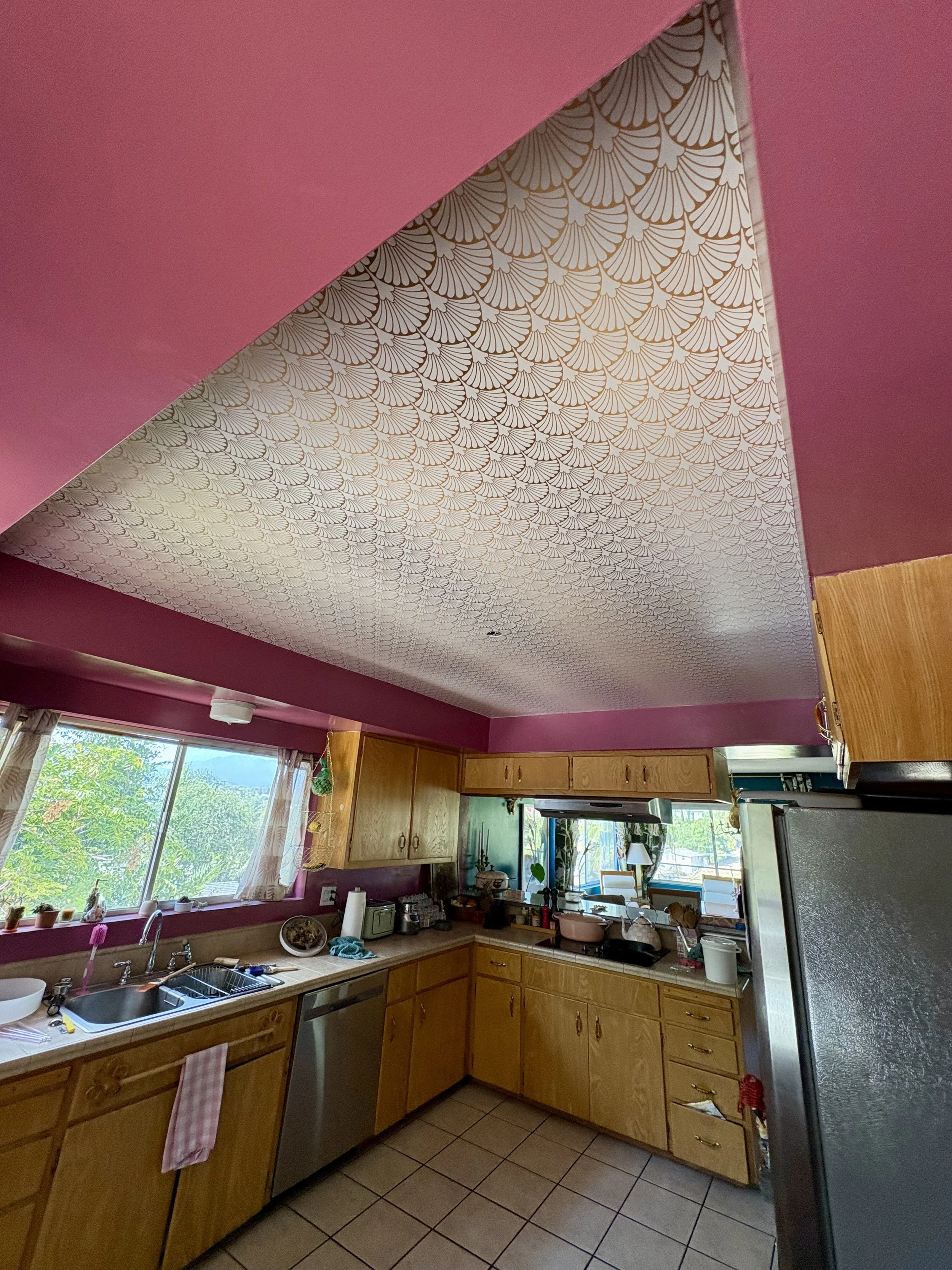 Spacious kitchen with wood cabinets, patterned wallpaper ceiling, tiled floor, large windows, sink, appliances, and stainless steel refrigerator
