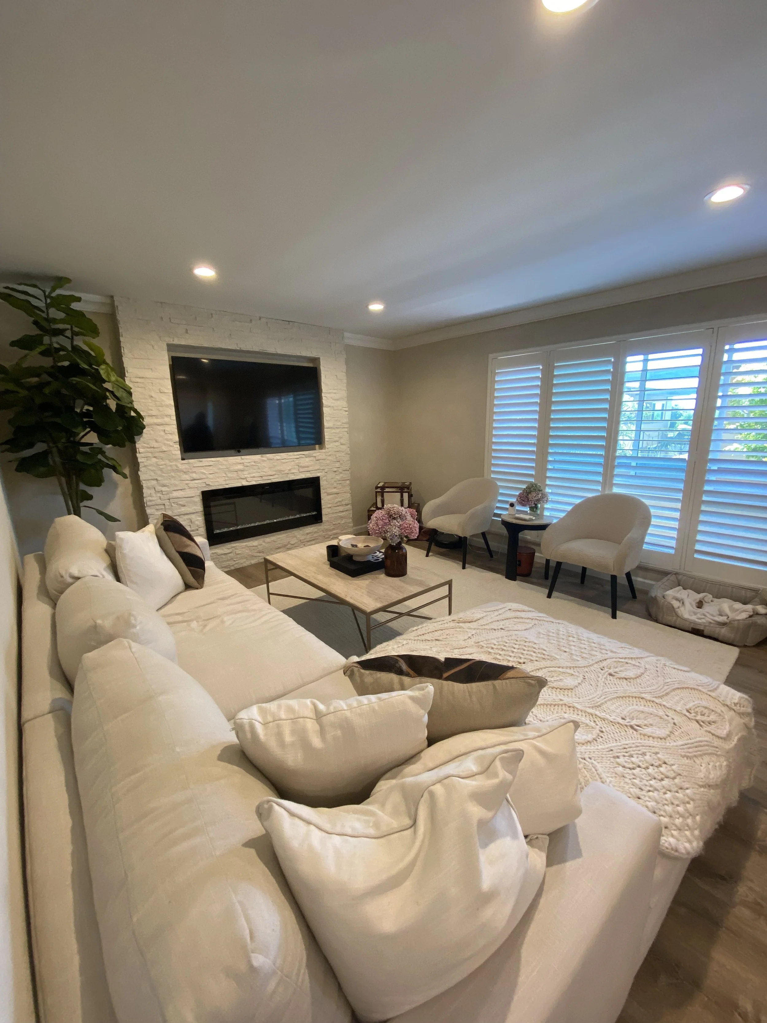 Bright modern living room with a white sectional sofa, stone fireplace, wall-mounted TV, armchairs, coffee table, houseplant, and plantation-shuttered windows.