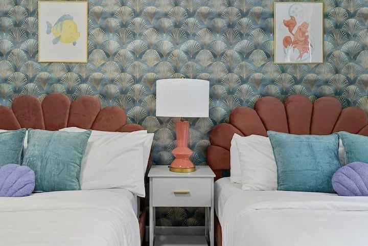 Colorful bedroom with twin beds, coral scalloped headboards, turquoise pillows, patterned wallpaper, framed artwork, and a coral table lamp