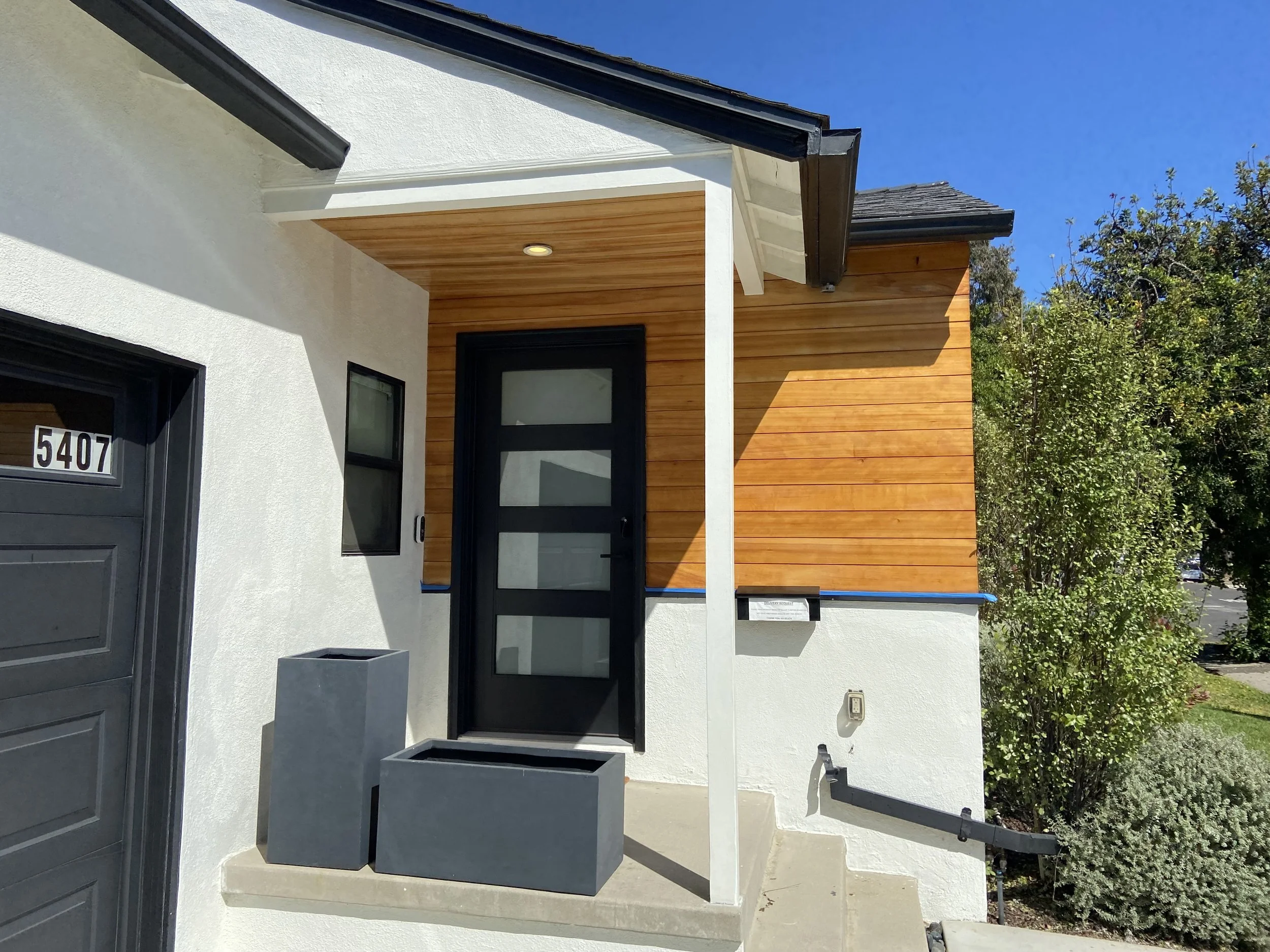 Modern home entrance with black glass-paneled door, warm wood siding, white stucco, planters, and house number 5407